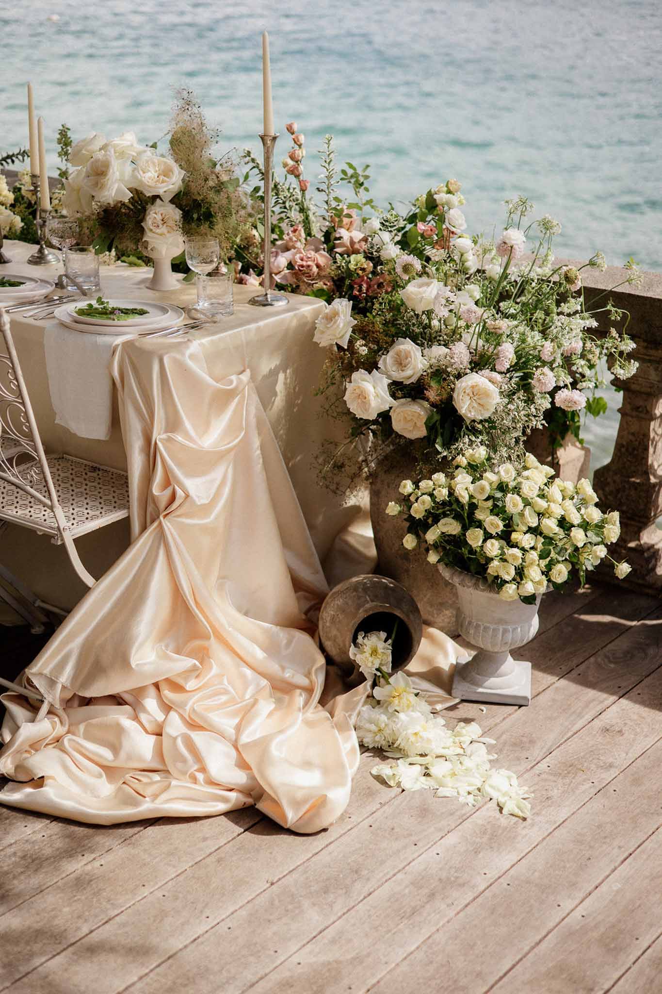 Waterfront tablescape with champagne satin cloth ivory roses and overflowing stone urn arrangement