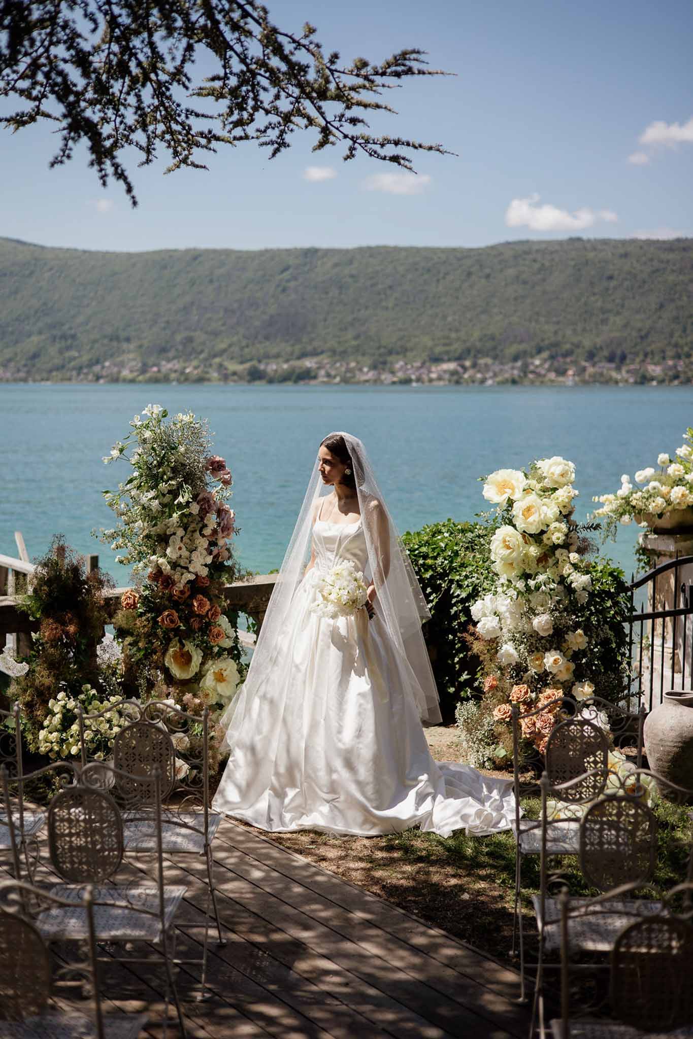Bride in white satin ball gown and cathedral veil on terrace overlooking turquoise lake with floral arrangements