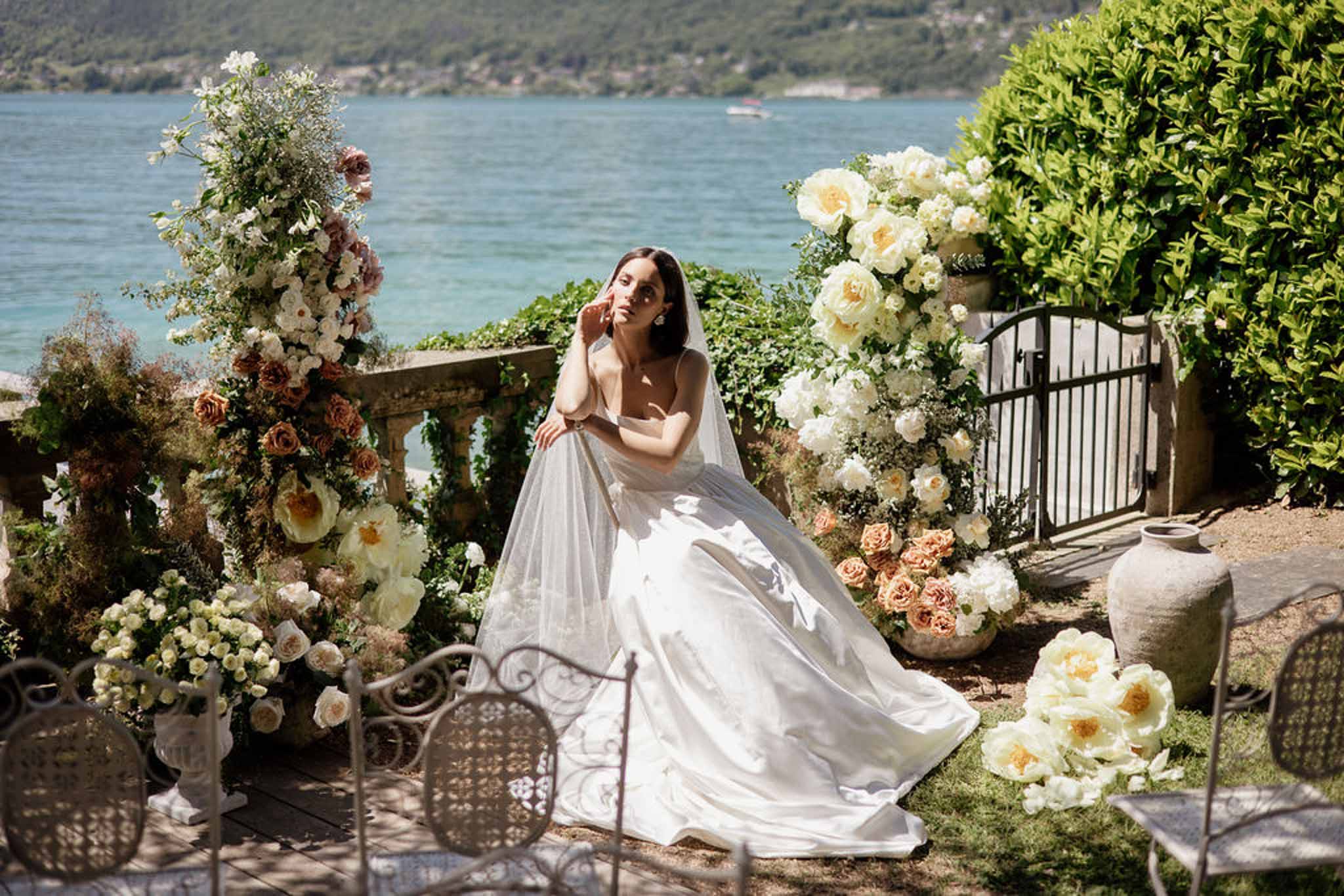Bride seated on lakeside terrace with cathedral veil amid peach and terracotta floral urns and columns