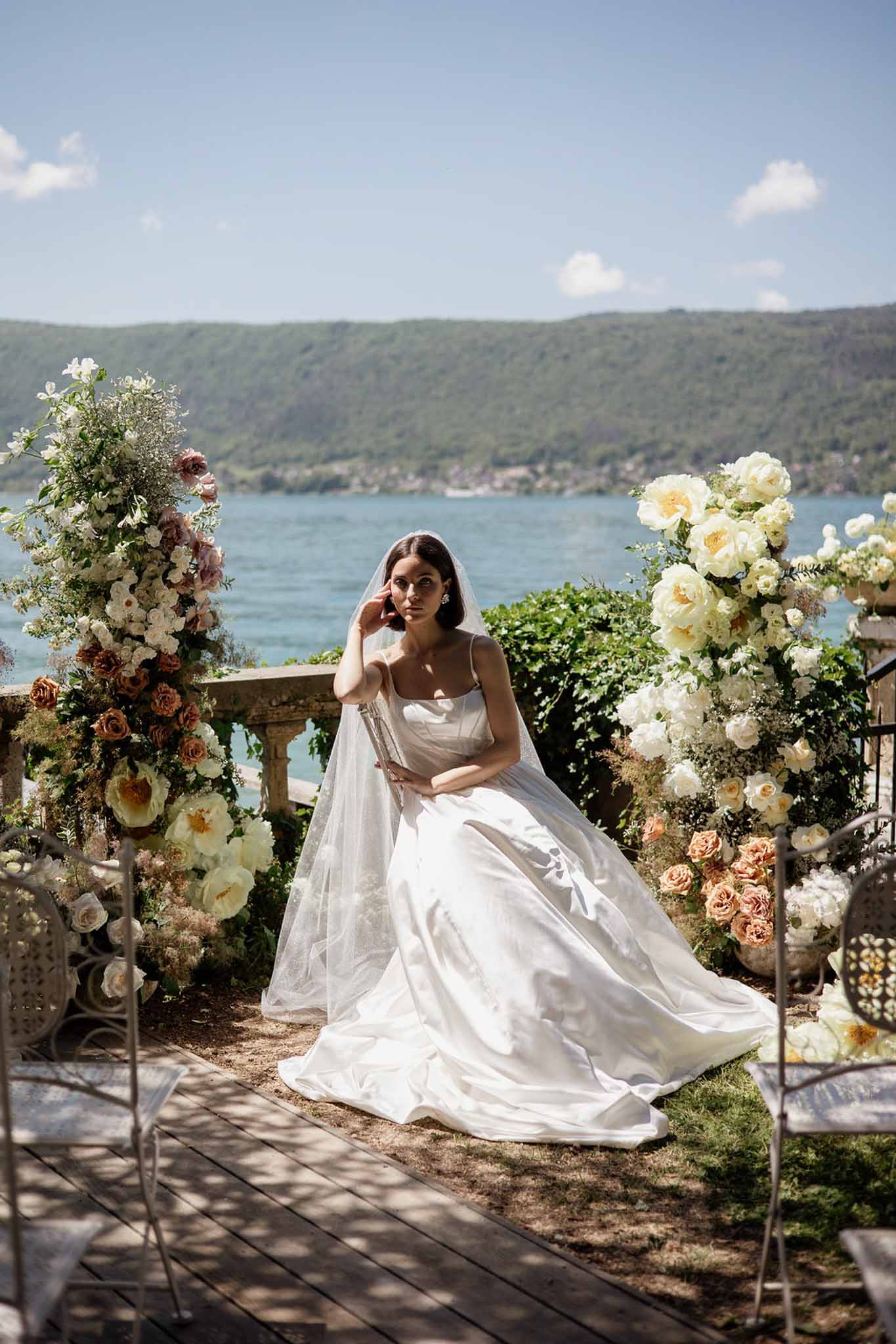 Bride seated on lakeside terrace with peach and terracotta rose arrangements on stone balustrade