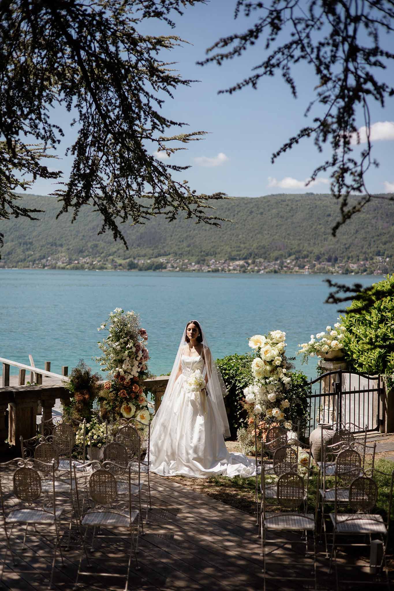 Bride at lakeside altar with terracotta and cream floral towers beside wrought-iron chairs and turquoise lake