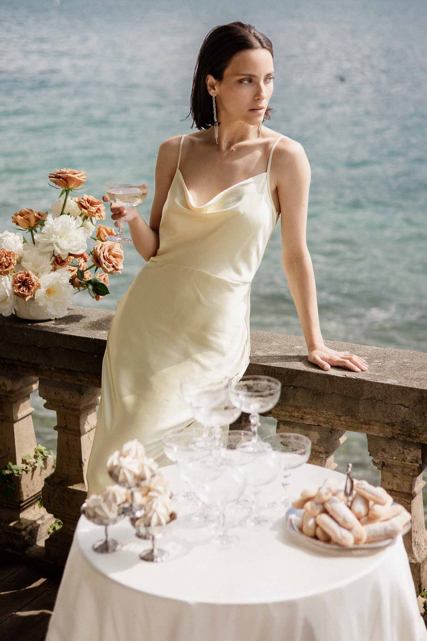 Bride in champagne slip dress at waterfront balustrade with champagne tower and terracotta roses
