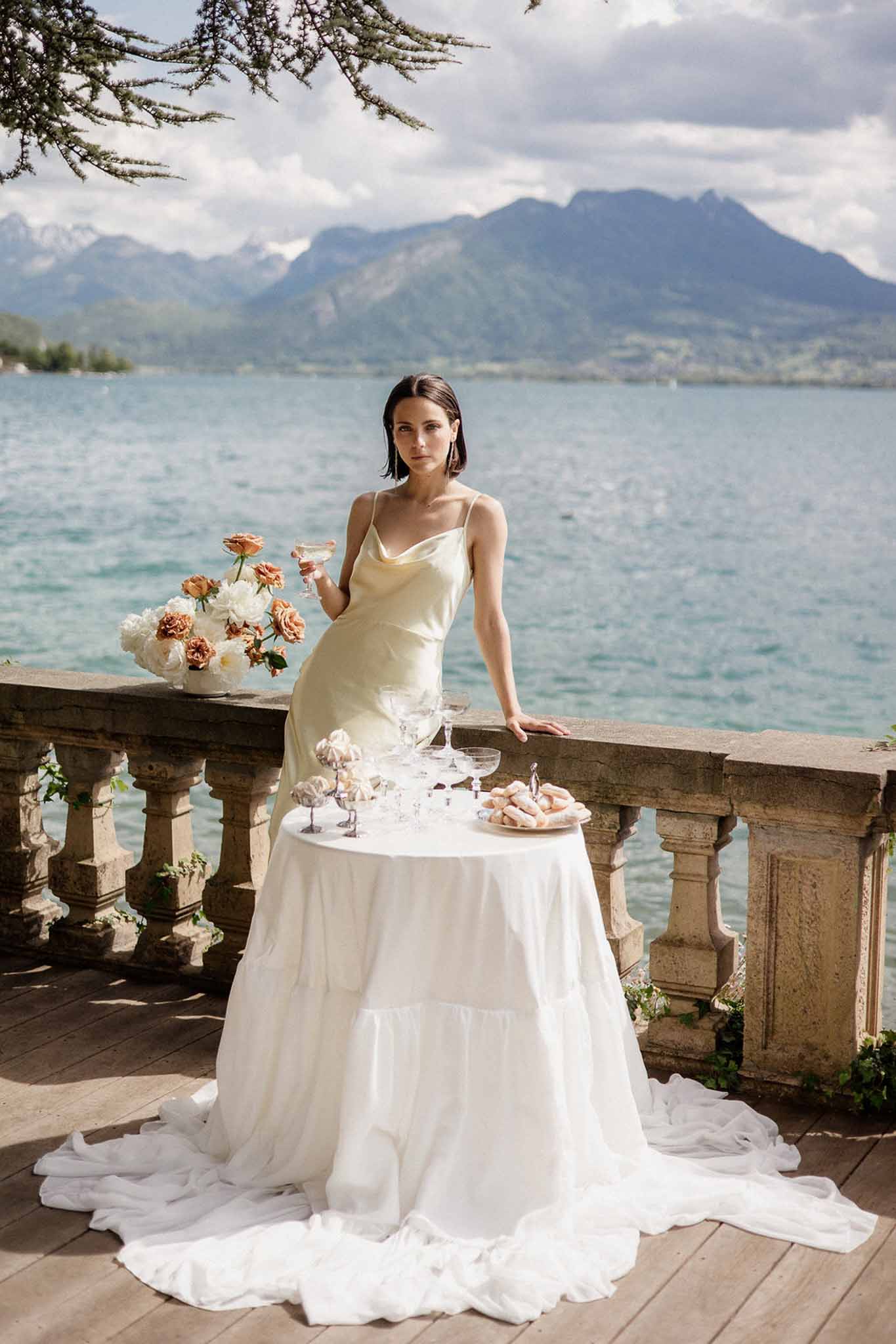 Bride in champagne silk slip dress with champagne coupe on lakeside terrace with terracotta roses and alpine mountains