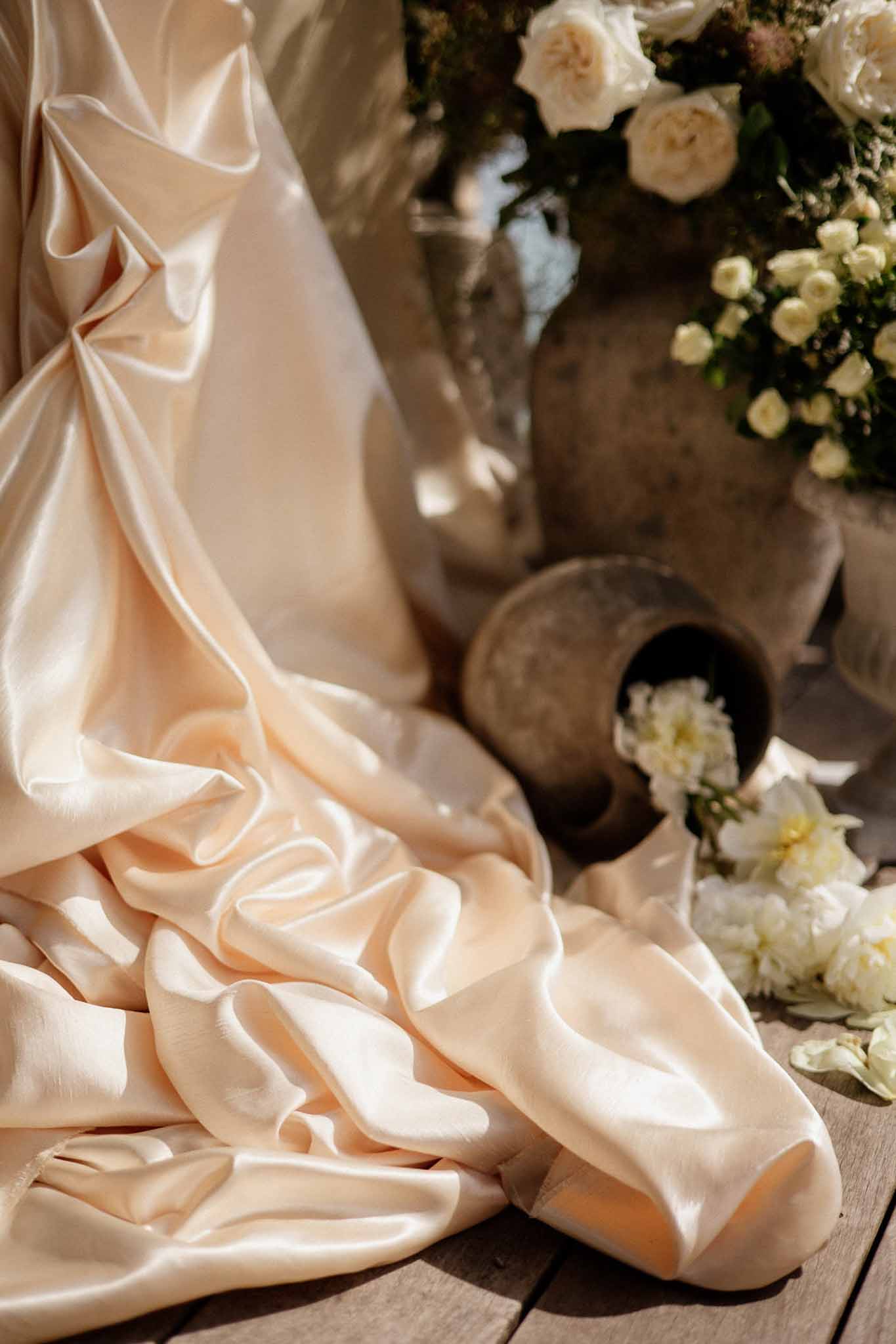 Blush-champagne satin bridal gown train beside stone vessels holding cream roses and white peonies on wooden surface