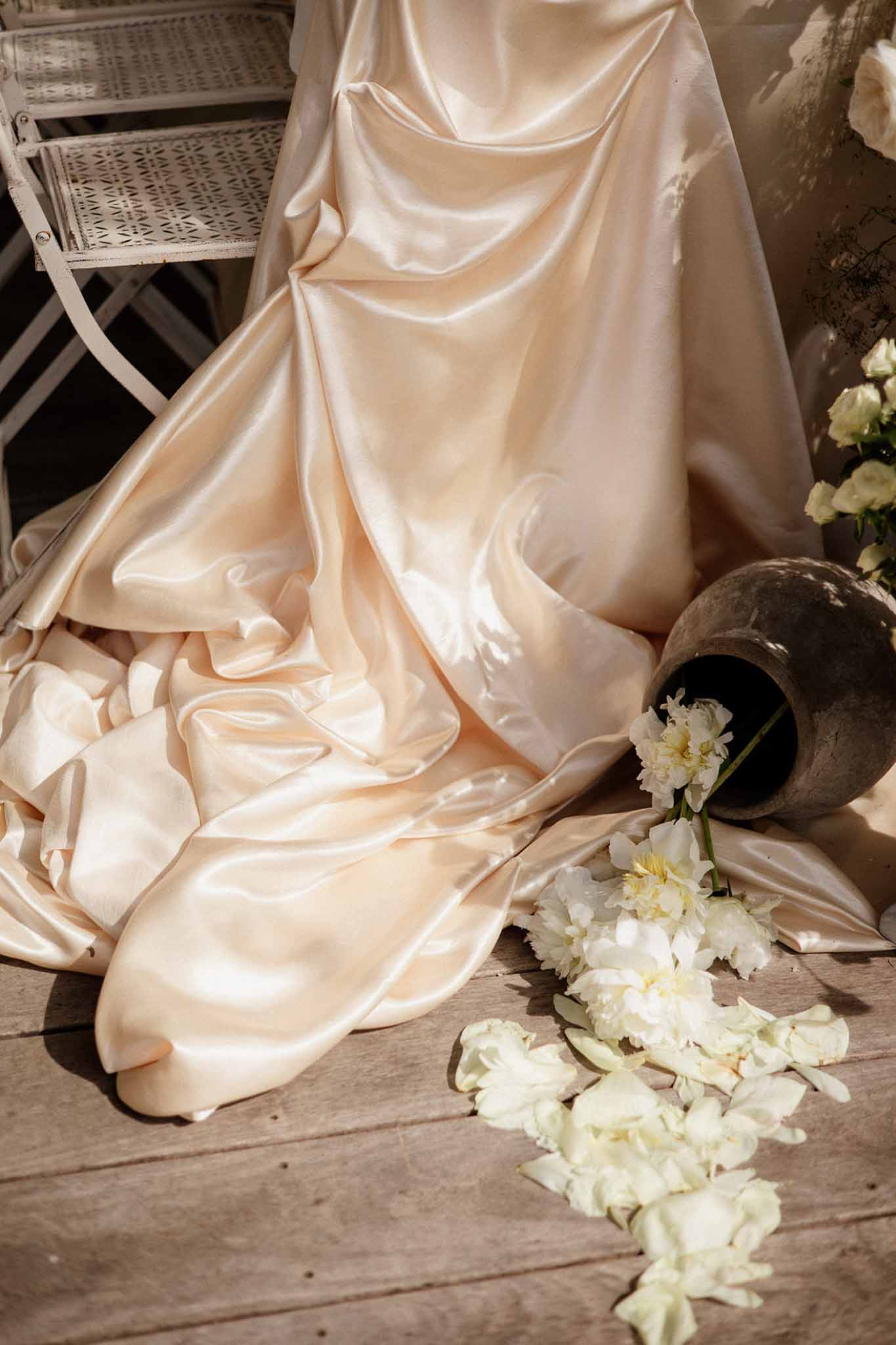 Close-up of champagne-blush satin wedding dress train with white peony blooms and scattered petals on wooden floor