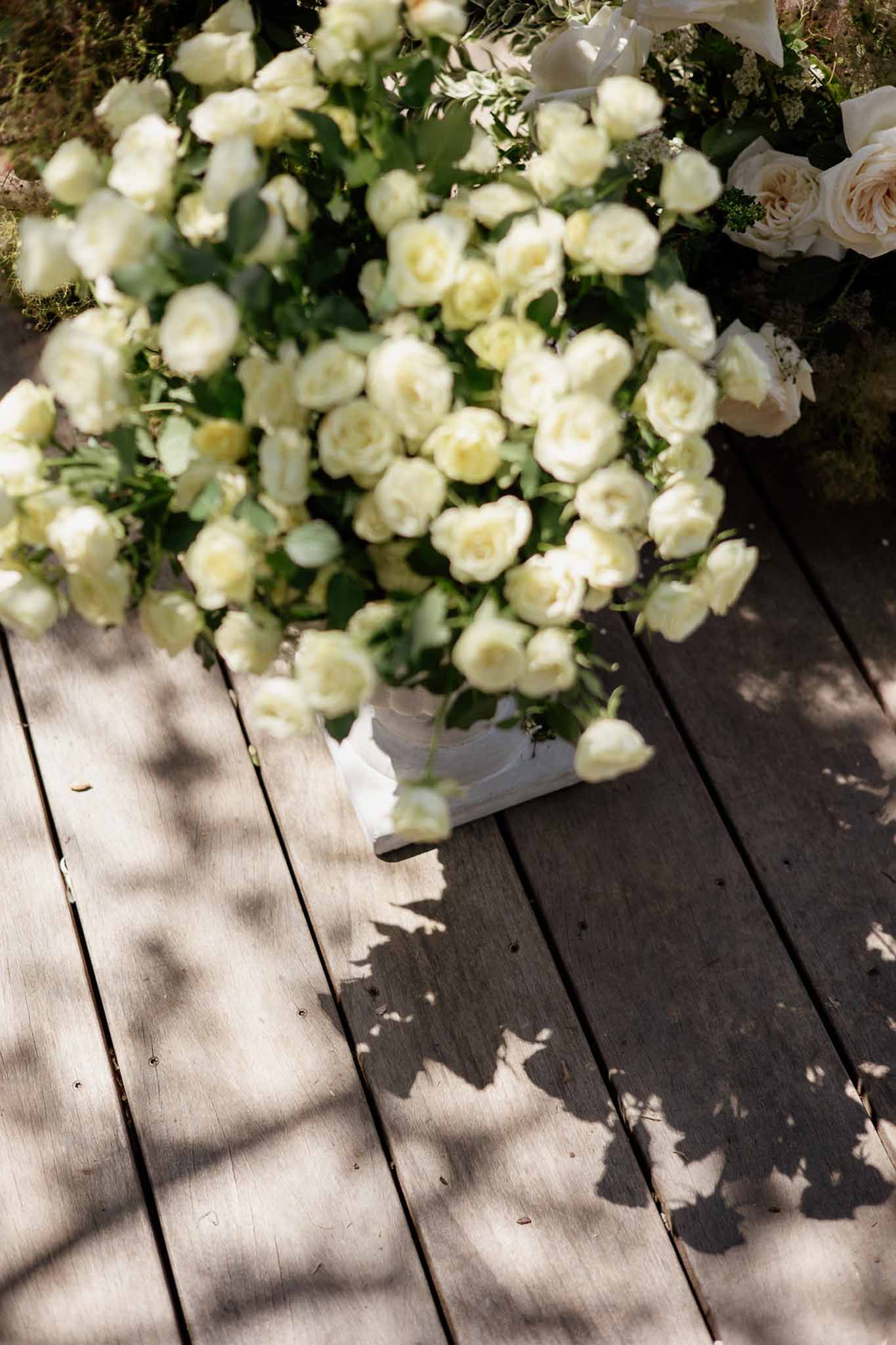 Cream and pale yellow spray rose arrangements in white containers on weathered wooden deck for ceremony decor