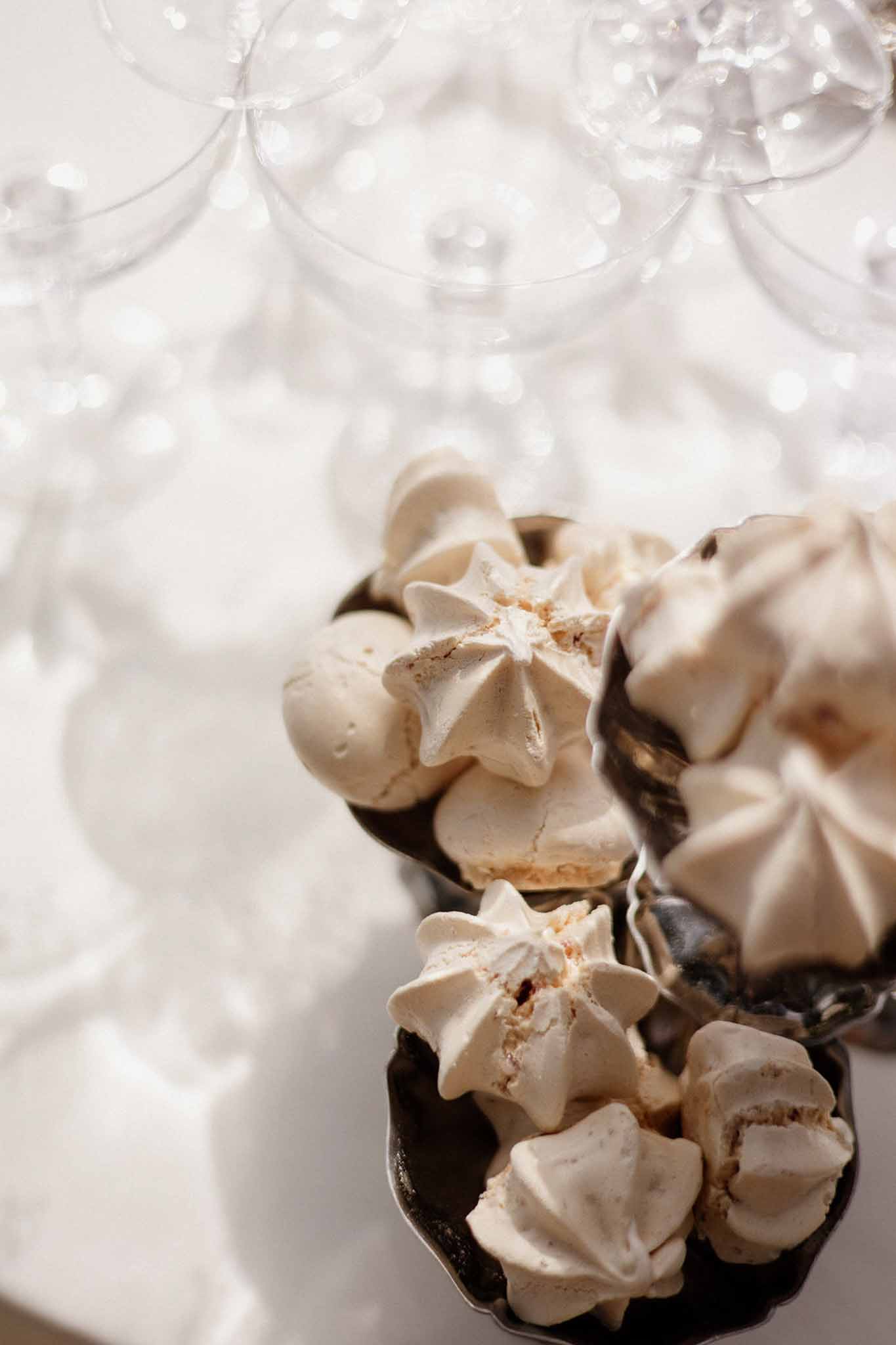 Ivory meringue kisses piled in dark metal compotes on a white linen table with champagne coupes behind