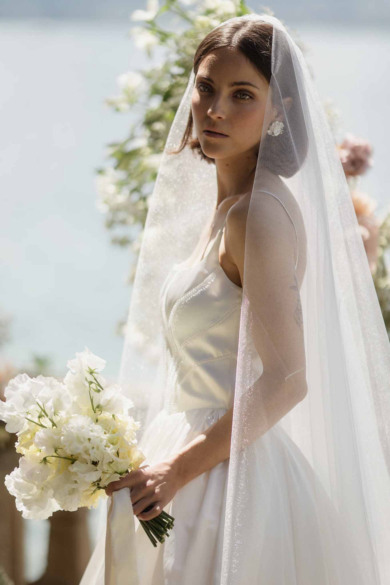 Bride in ivory satin slip gown and embellished veil holding white sweet pea bouquet by waterside