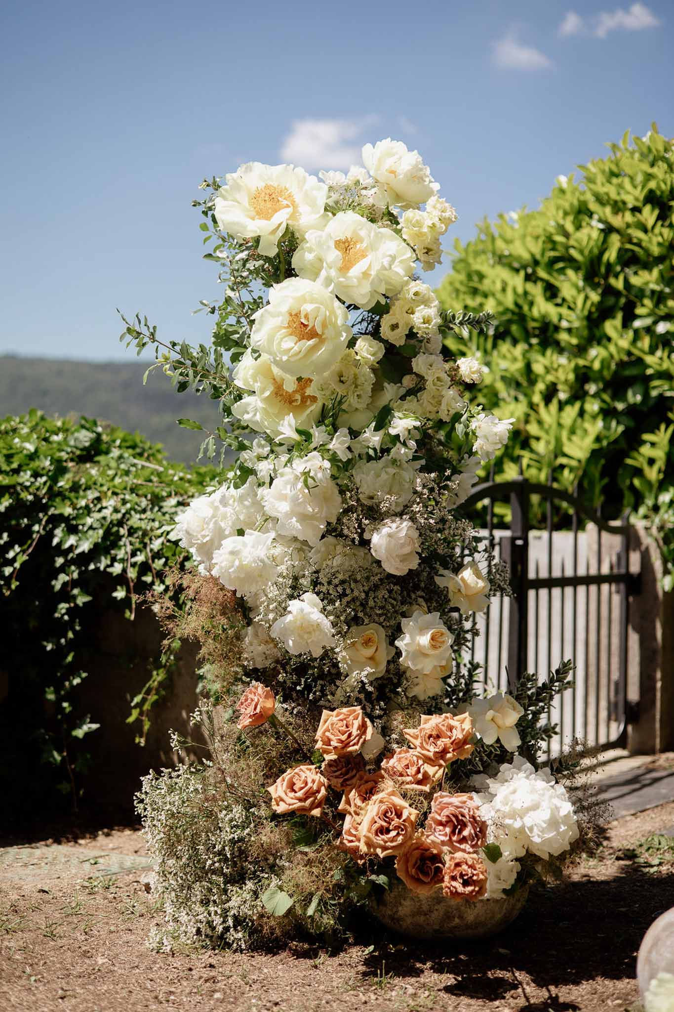 Tall floral column arrangement with terracotta roses, cream peonies, and trailing greenery at outdoor ceremony entrance