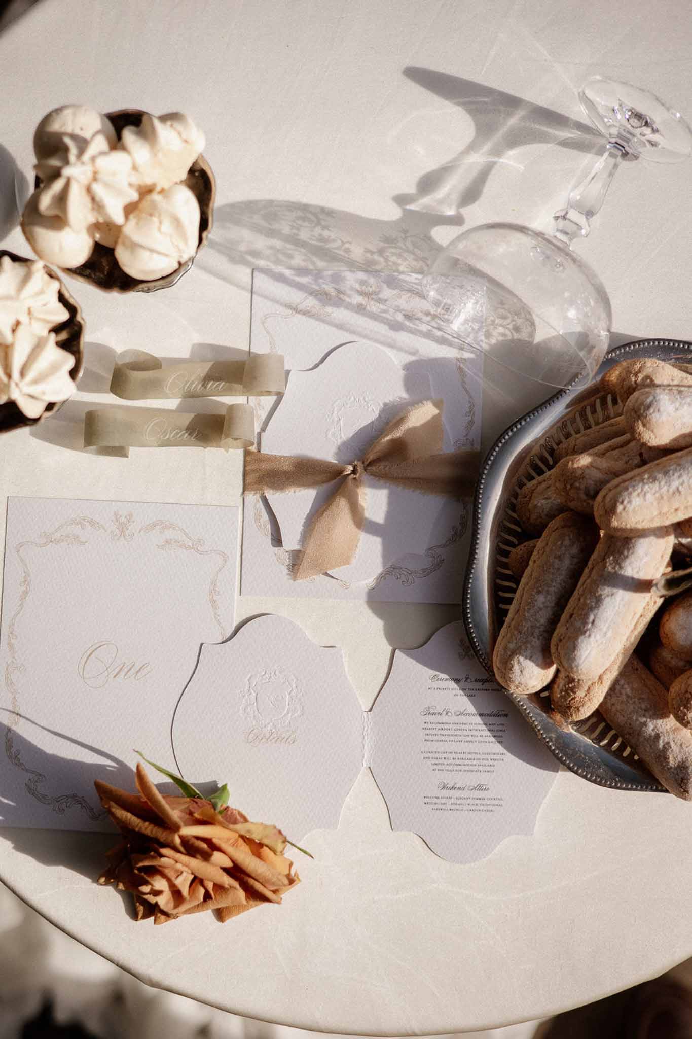 Flat lay with gold-bordered stationery sage ribbon place cards crystal glass ladyfingers and dried amber rose