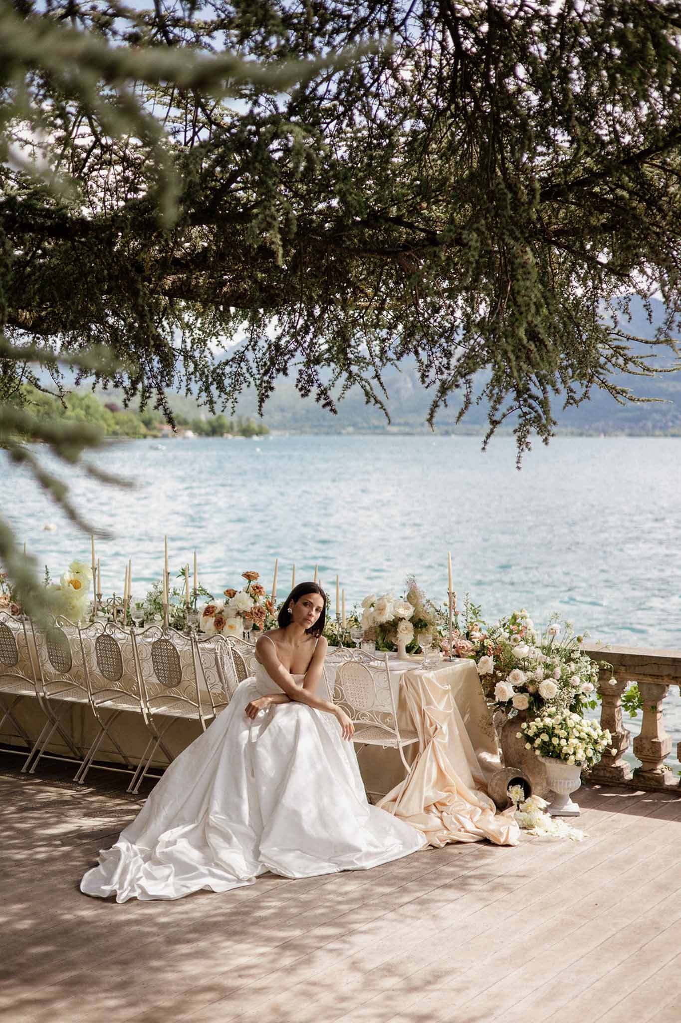 Bride in white satin ballgown seated on lakeside terrace beside a styled reception table with cream and apricot florals