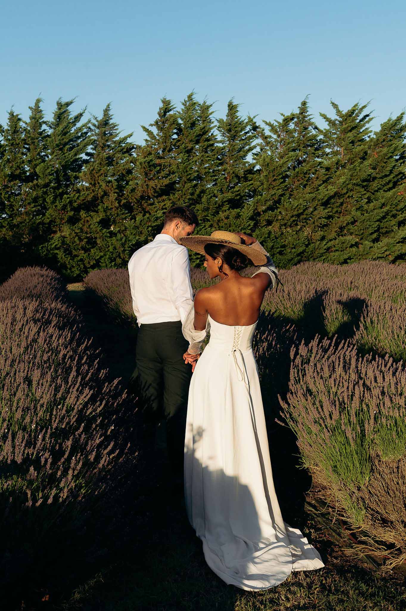 Rear view of bride and groom walking through lavender fields at golden hour, Bastide des Barattes