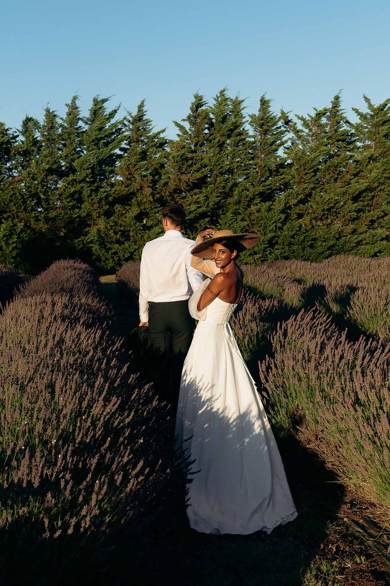 Bride and groom walking through lavender fields at golden hour, bride wearing wide-brim hat, Bastide des Barattes