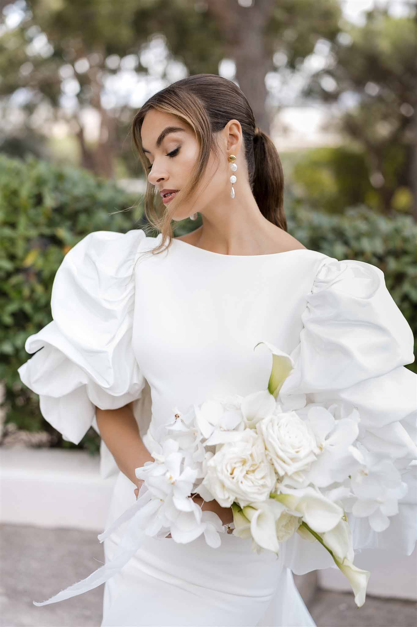 Close-up bride in bateau-neck gown with voluminous puff sleeves pearl earrings and all-white rose bouquet