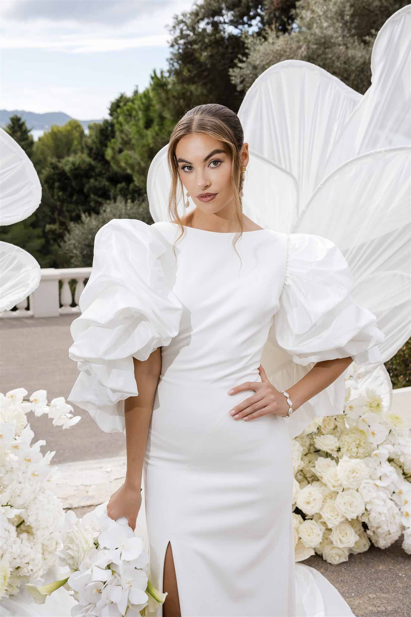 Bride in white crepe gown with puff sleeves holding orchid bouquet surrounded by sculptural white floral decor