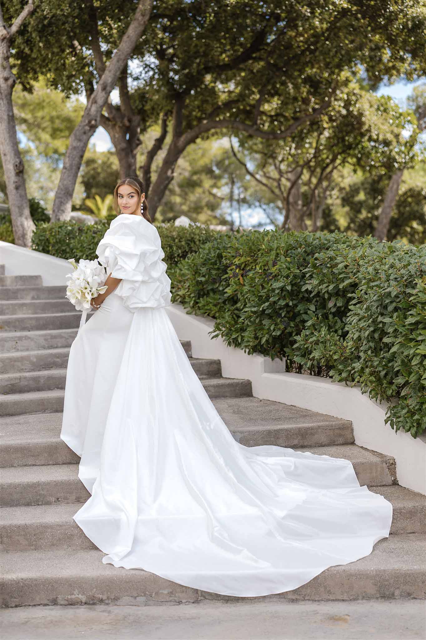 Bride looking over shoulder on stone staircase wearing ruffled off-shoulder gown with cathedral train