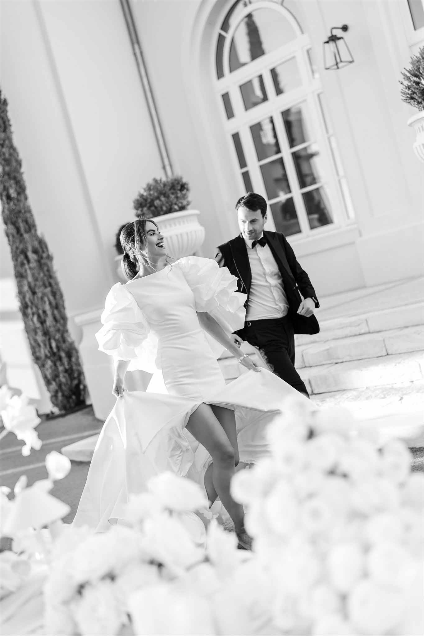 Black and white couple running and laughing before classical arched facade with topiary and white florals