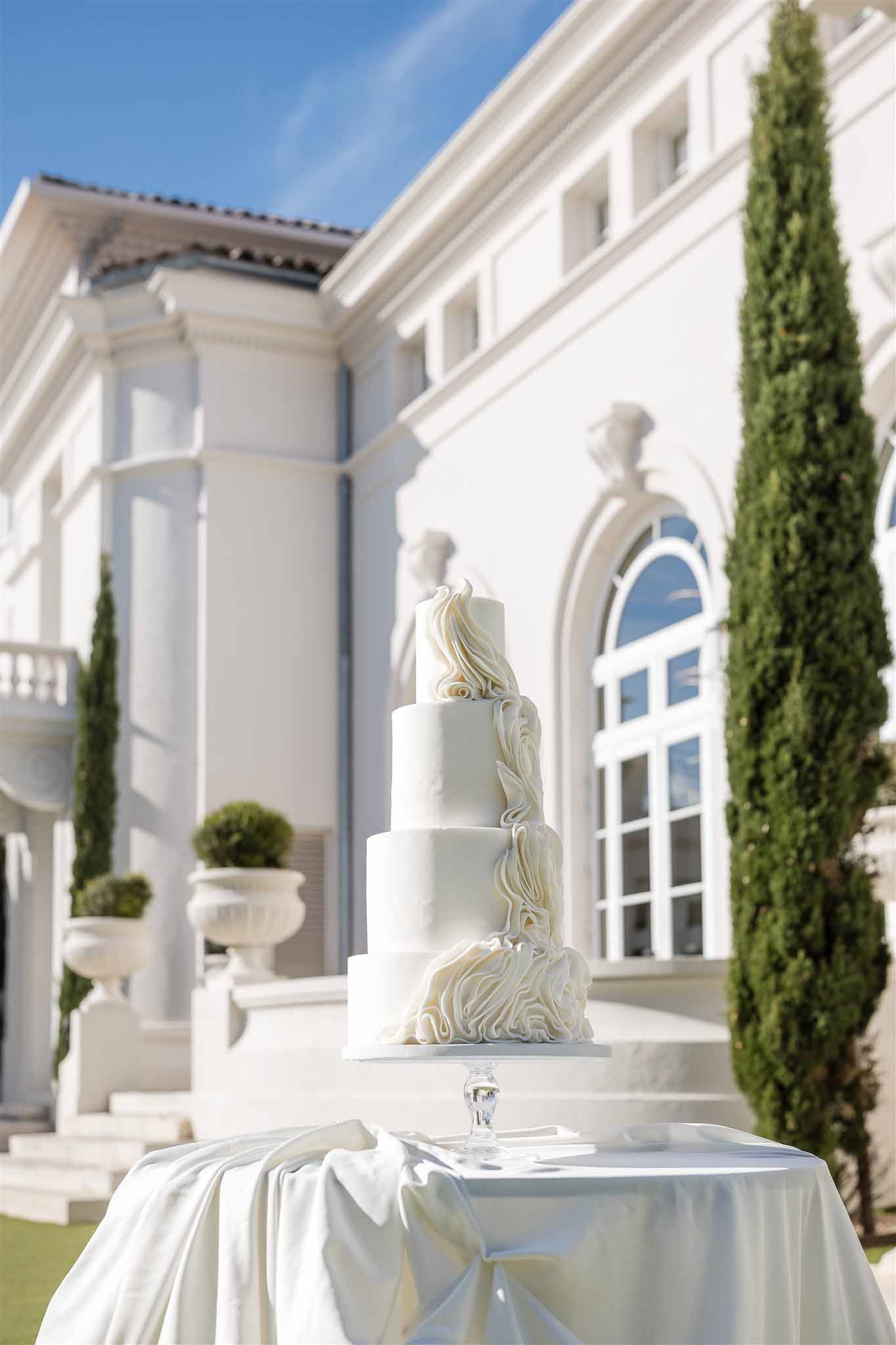 Four-tier white ruffle wedding cake on crystal stand before neoclassical villa with cypress trees and stone urns