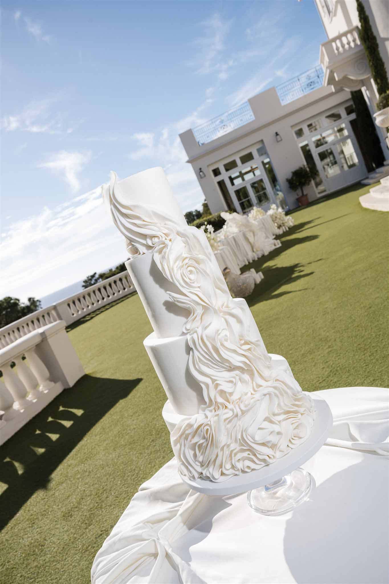 Multi-tier white ruffled rosette cake on glass stand before white villa with reception tables on lawn