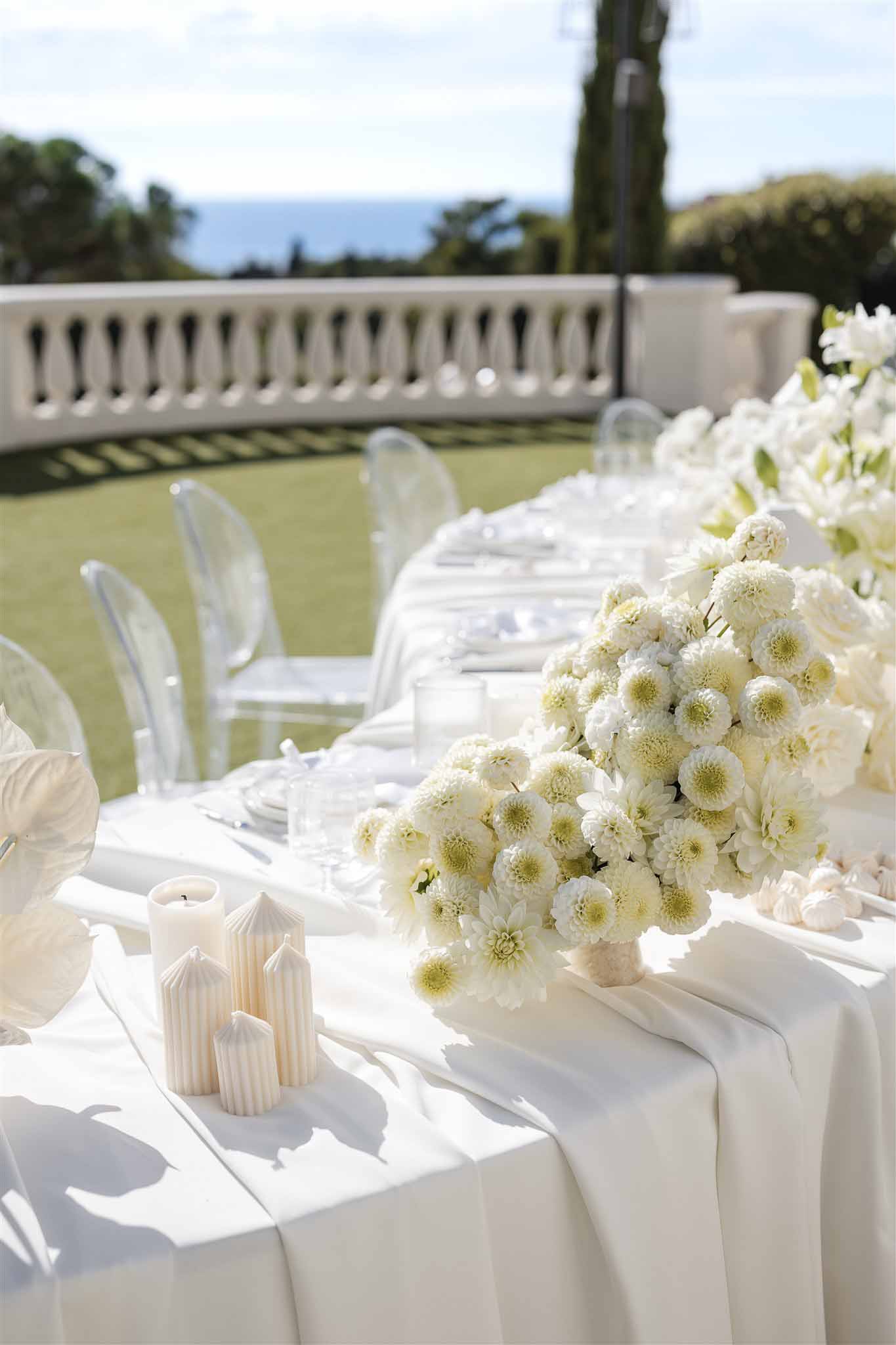 All-white reception table with chrysanthemum dome centerpiece satin runner ribbed pillar candles and ghost chairs