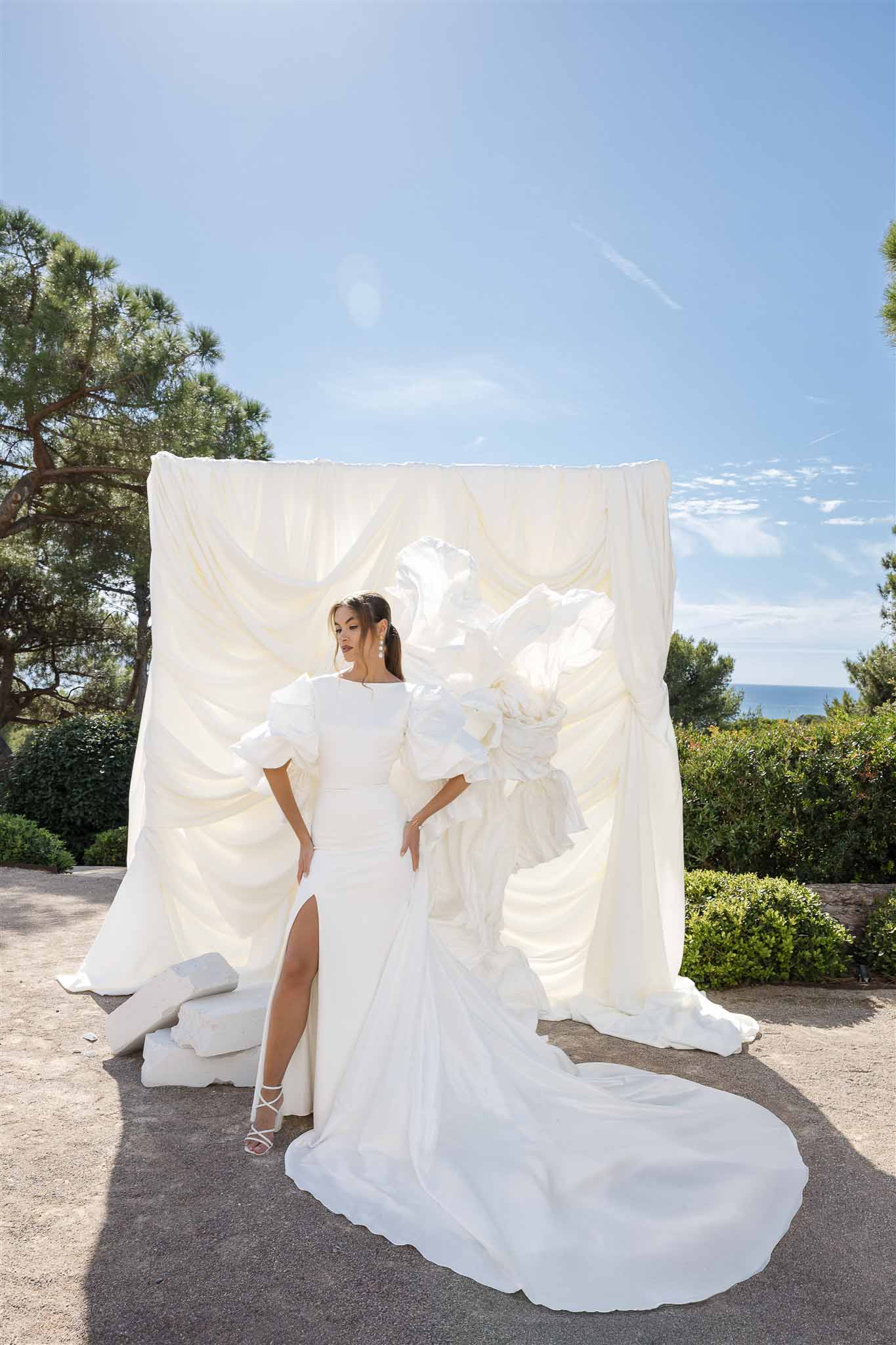 Bride in puff-sleeve gown with thigh slit and train before ivory draped backdrop with organza sculpture