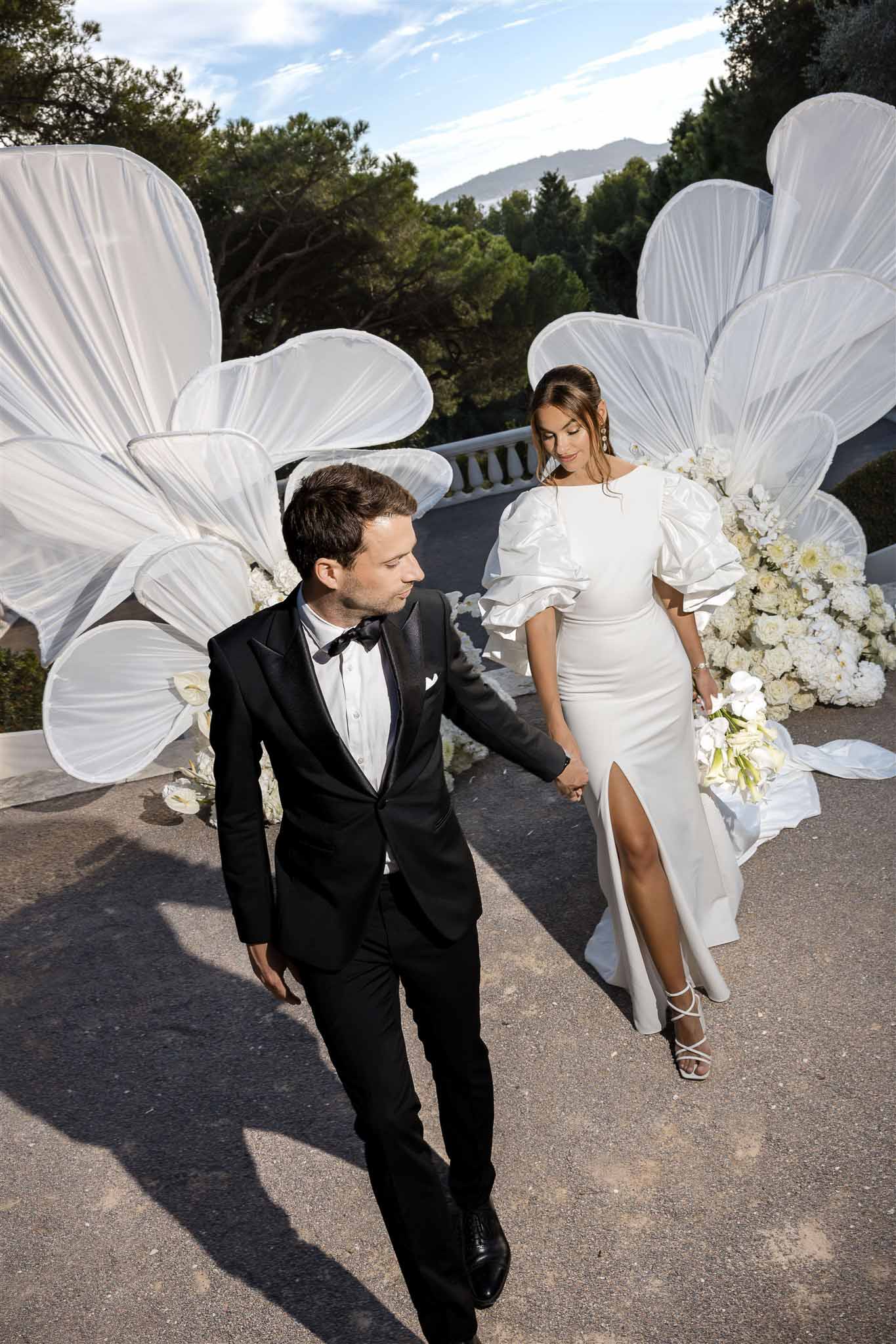 Couple walking past oversized white fabric flower sculptures with calla lily bouquet