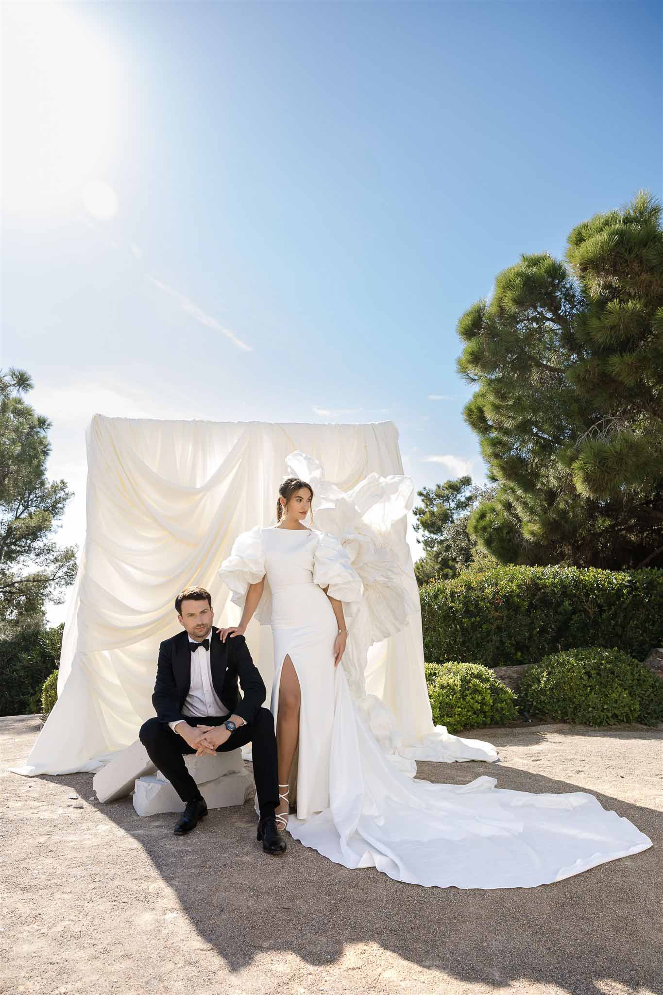 Bride standing in ruffle-sleeve gown with seated groom in tuxedo before large draped ivory fabric installation