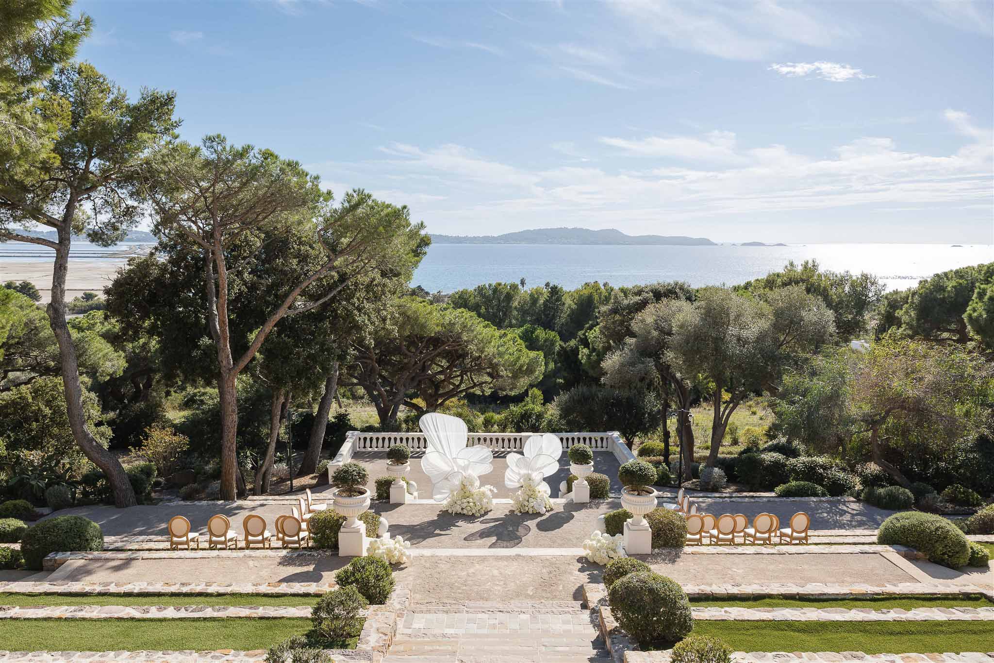 Aerial view of outdoor ceremony setup on coastal terrace with white petal backdrop, gold chairs, and Mediterranean Sea beyond