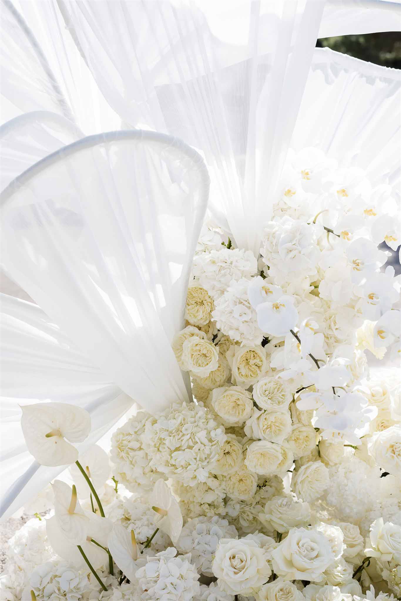 All-white ceremony arch with garden roses, orchids, calla lilies, and sculptural acrylic fan elements