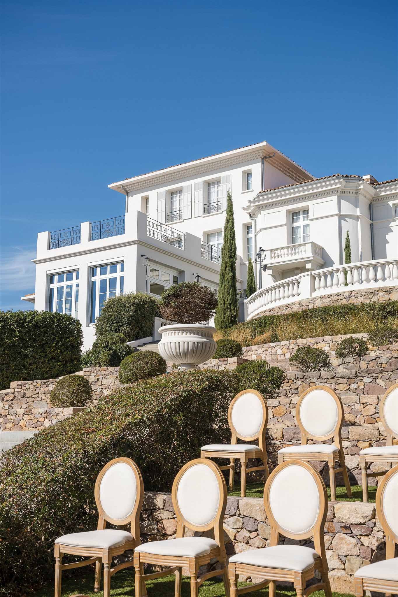 Gold Louis XVI chairs arranged on a lawn facing a white Provencal villa with wrought-iron balconies and stone staircase