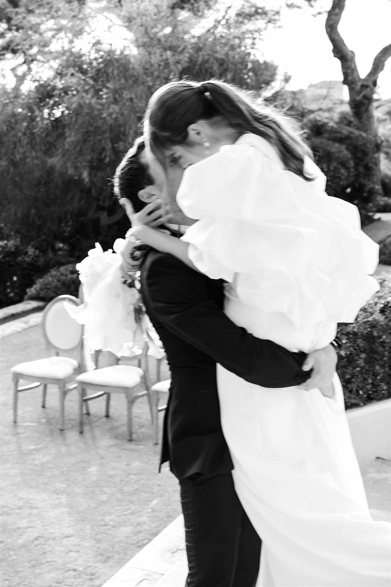 Black-and-white image of groom lifting bride for a kiss after outdoor ceremony with gold-framed chairs behind