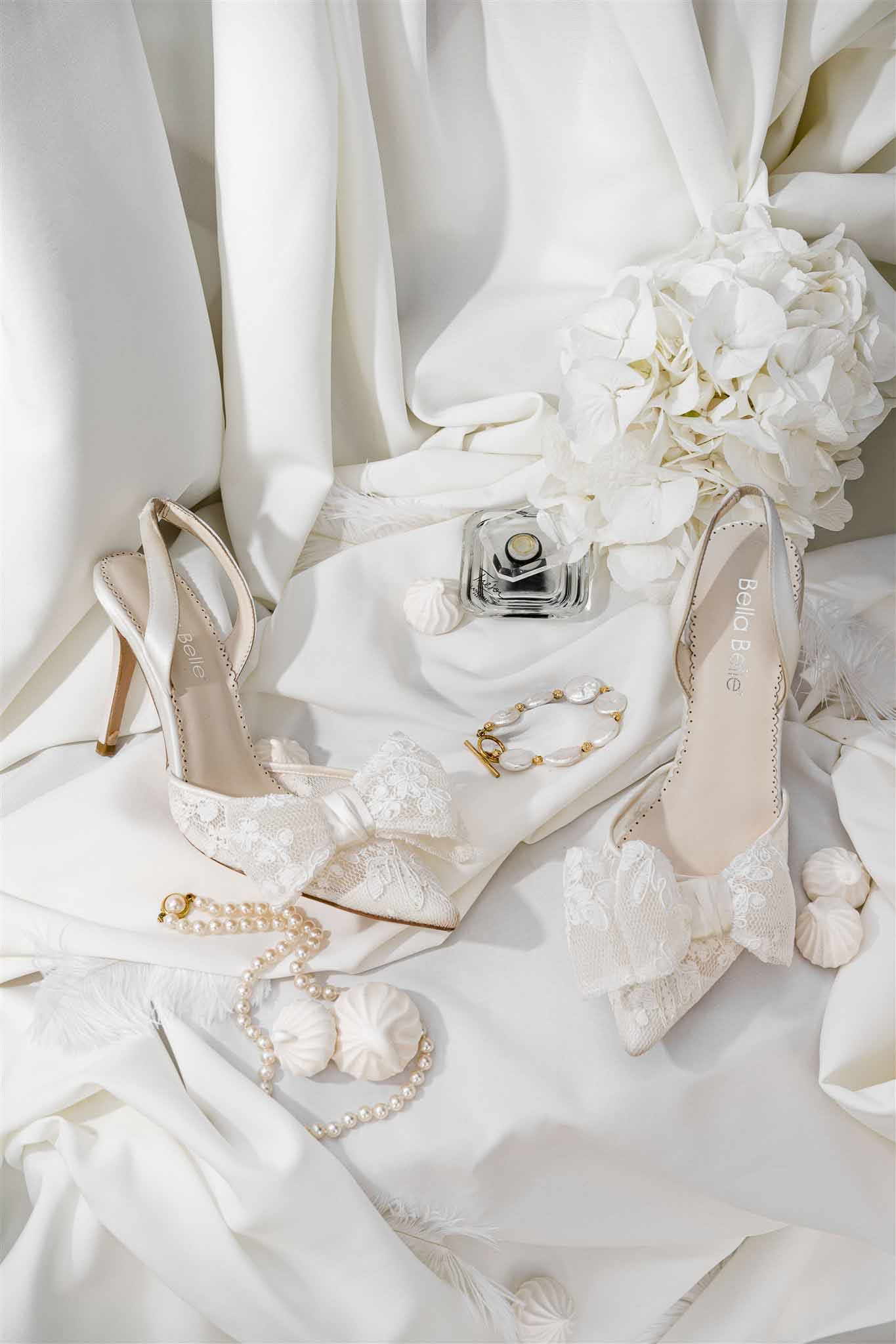 Bella Belle ivory satin heels with lace bows, pearl bracelets, perfume, and hydrangea on white satin