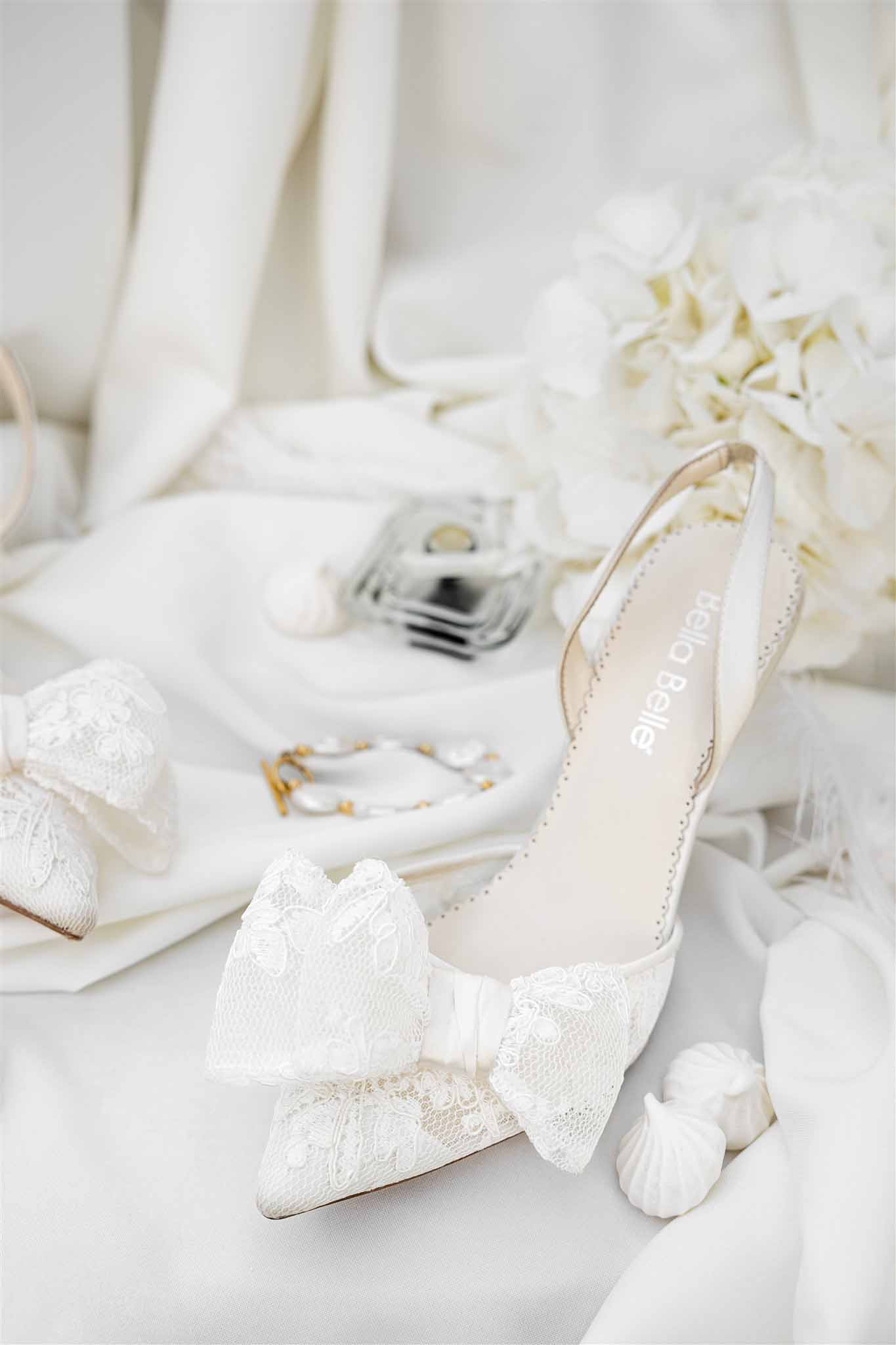 Bridal flat lay with ivory lace bow slingback heels, perfume bottle, gold bracelet, and Jordan almonds on satin