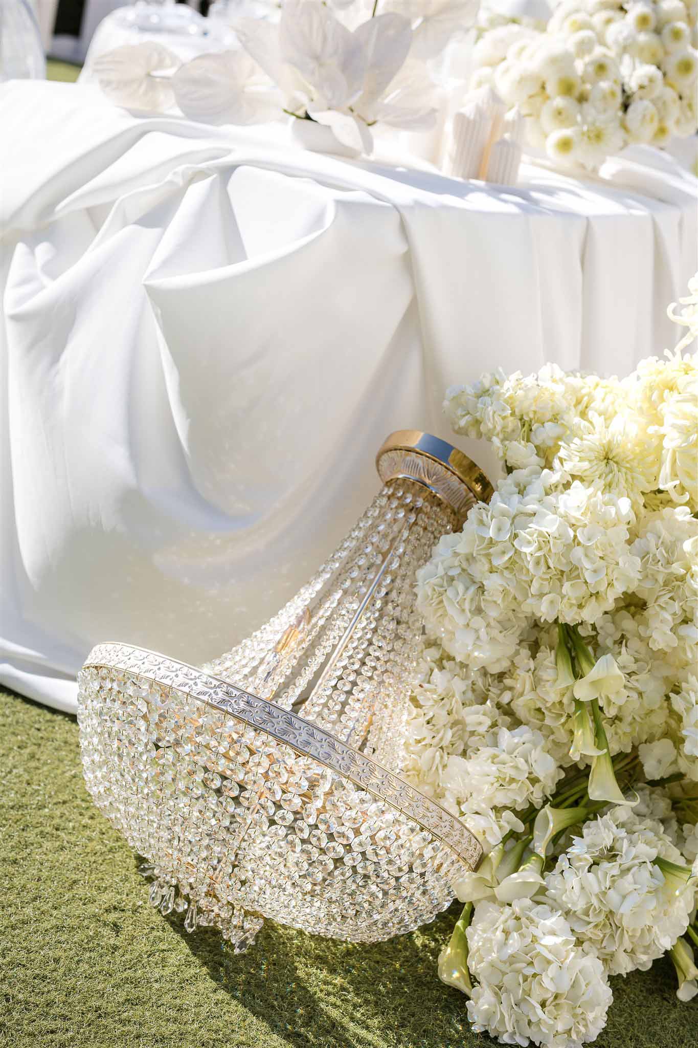 Crystal and gold chandelier on a lawn beside ivory hydrangeas and white calla lilies with reception table behind