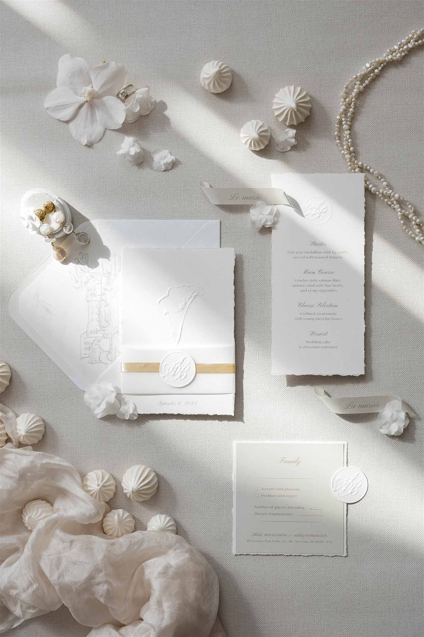 Embossed invitation suite with gold ribbon seal, pearl necklace, and meringues on grey linen