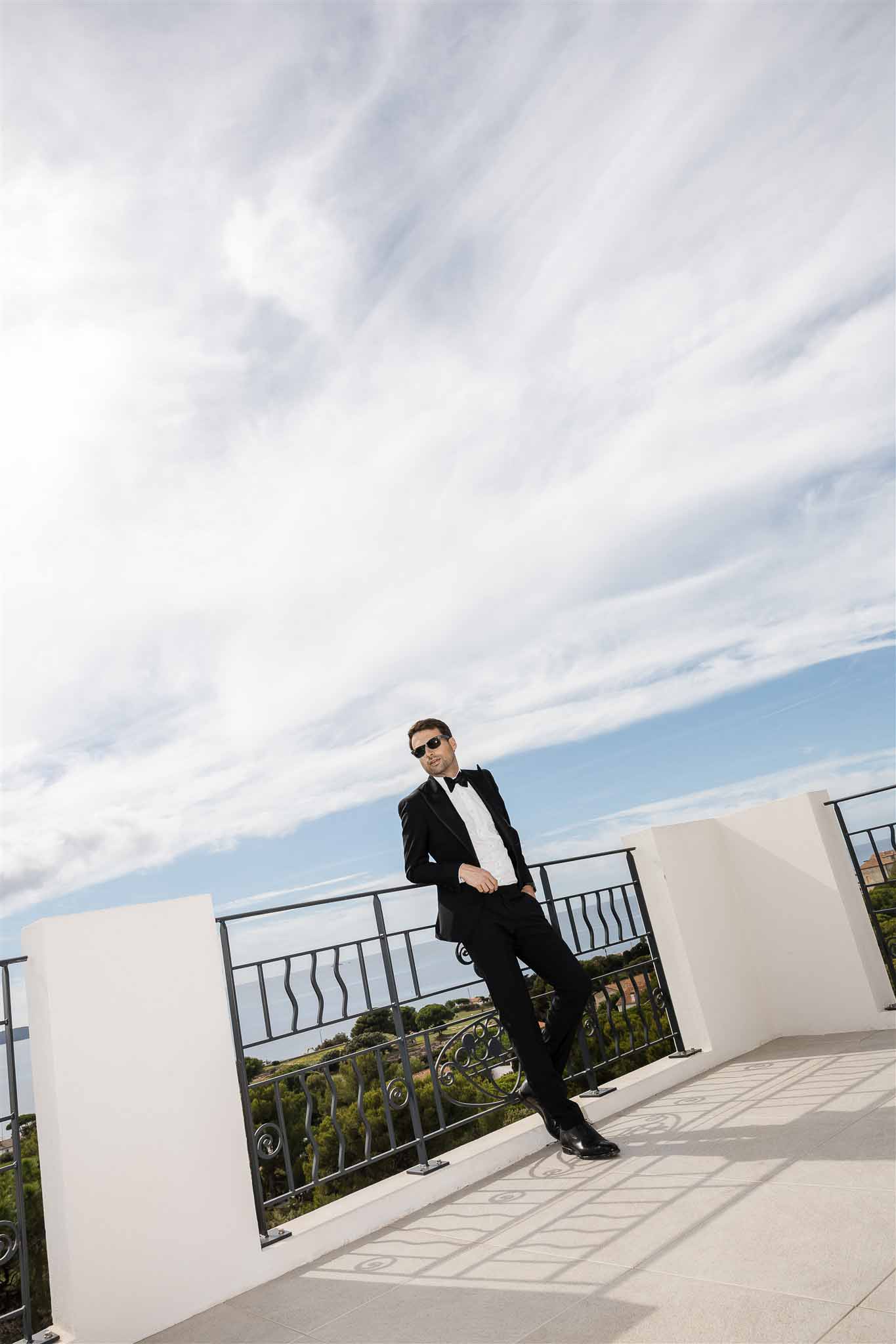 Groom in black tuxedo and sunglasses leaning against wrought-iron railing on a rooftop terrace