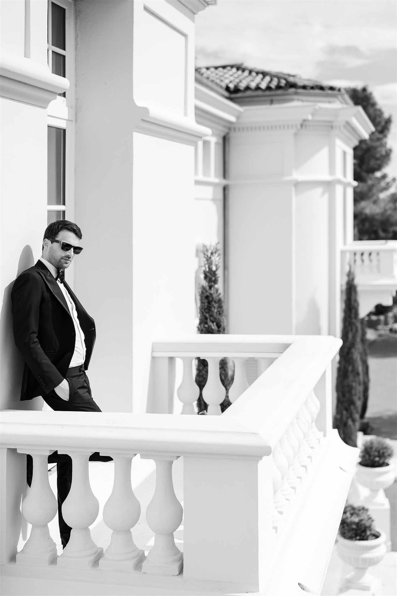 Black and white full-length portrait of groom in tuxedo leaning against classical white balustrade at formal venue