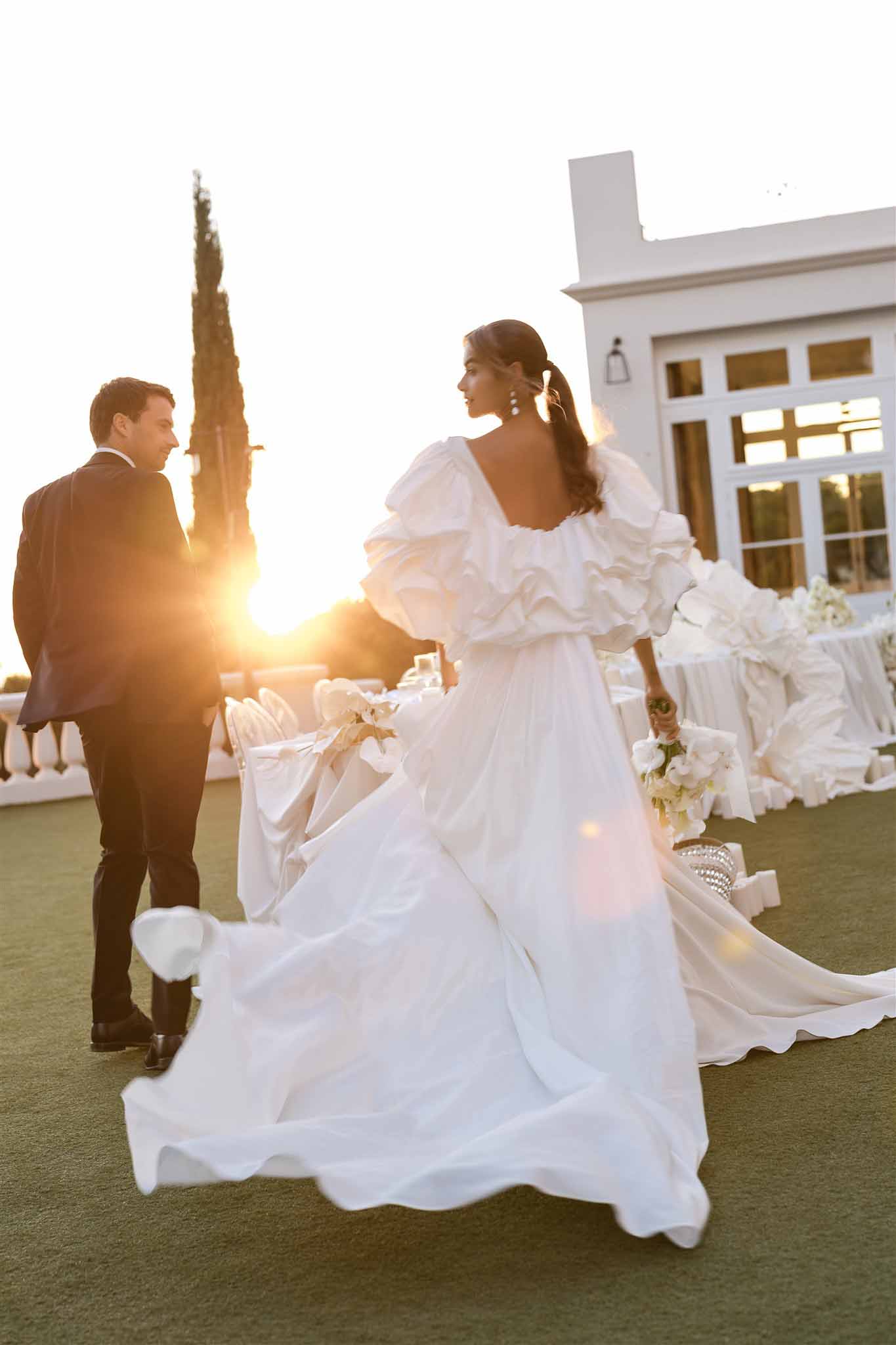 Bride in dramatic puff-sleeve ball gown and groom in navy suit at golden hour with white reception table behind