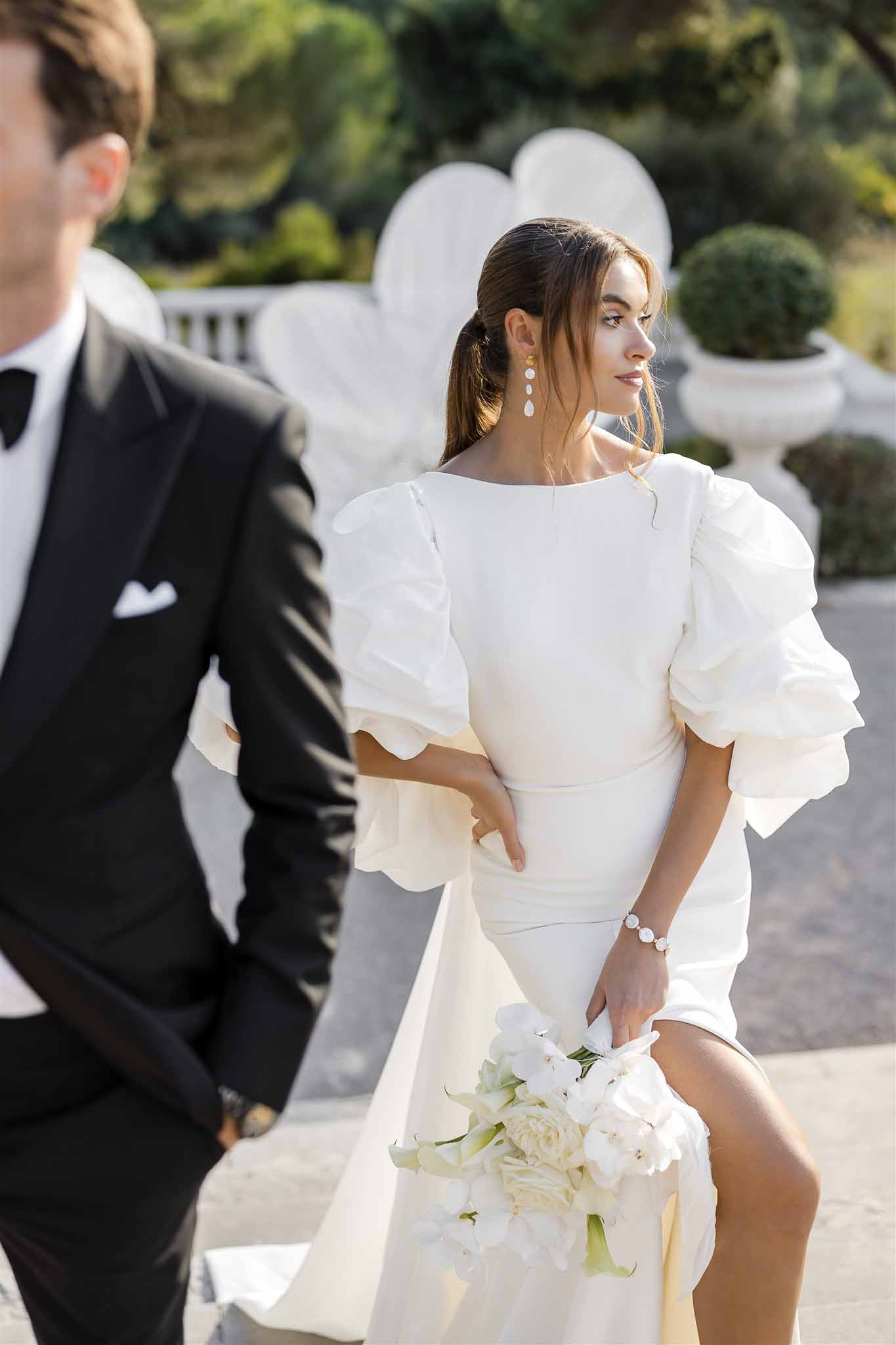 Bride in fitted gown with organza puff sleeves holding white orchid bouquet beside groom on terrace