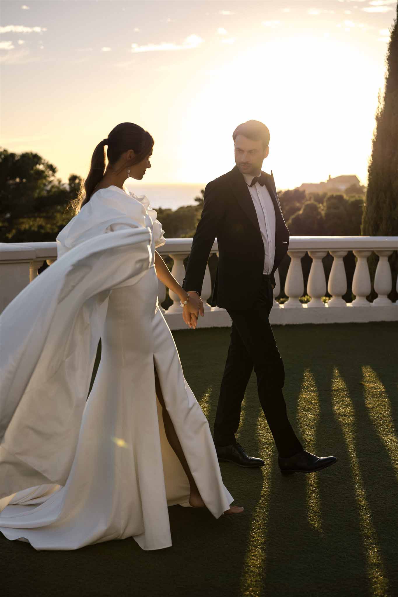 Couple walking on balustrade terrace at golden hour with dramatic cape gown and long diagonal shadows