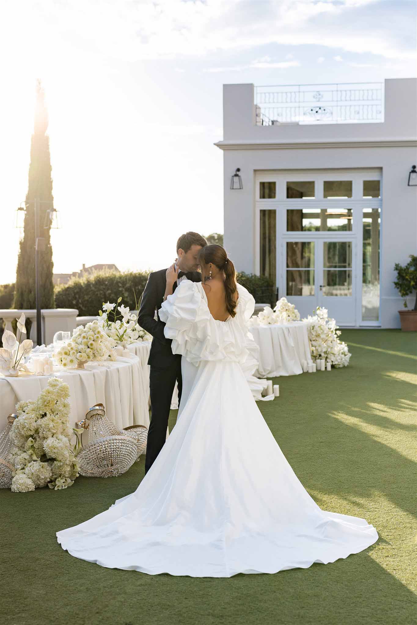 Groom kissing bride hand at golden hour with all-white hydrangea reception tables and crystal chandeliers