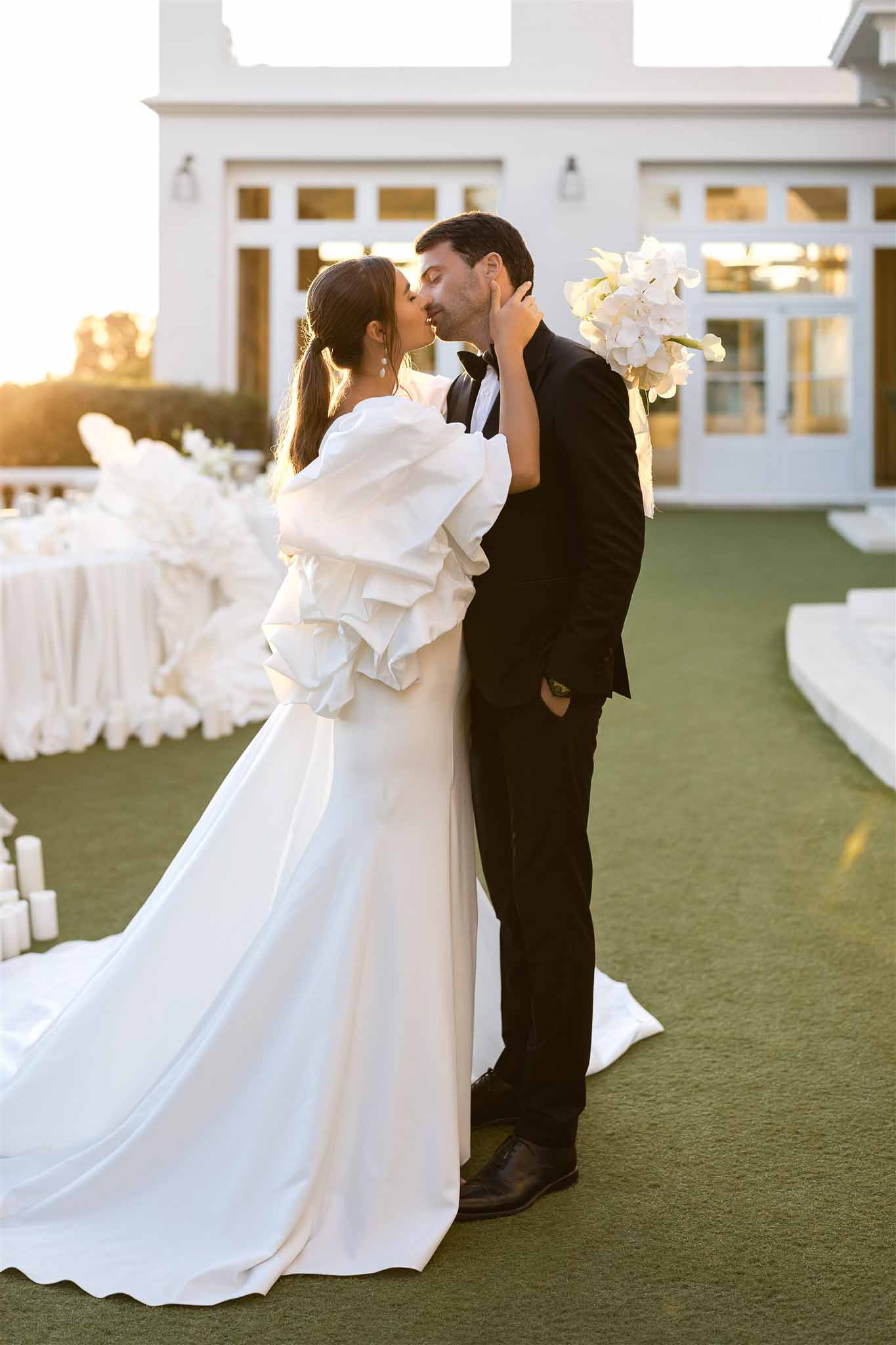 Couple kissing at golden hour on lawn before white building with orchid bouquet and reception table in background