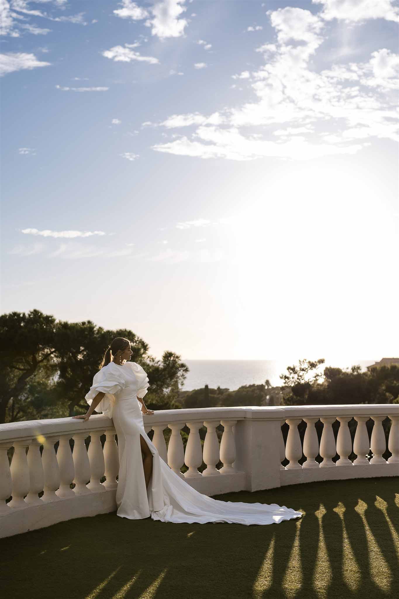 Bride in fitted gown with puff sleeves and long train poses on stone balustrade terrace at sunset