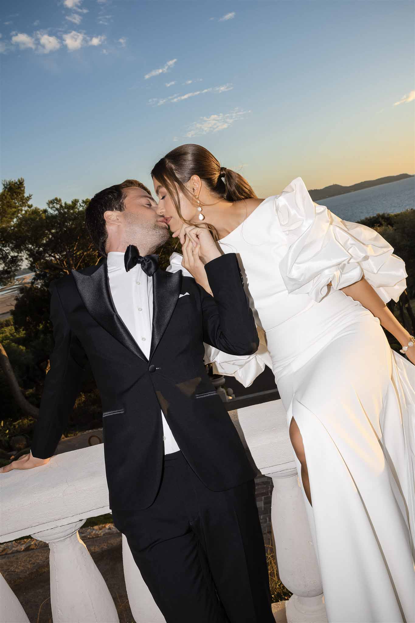 Couple kissing on balcony overlooking sea at golden hour with coastal landscape behind