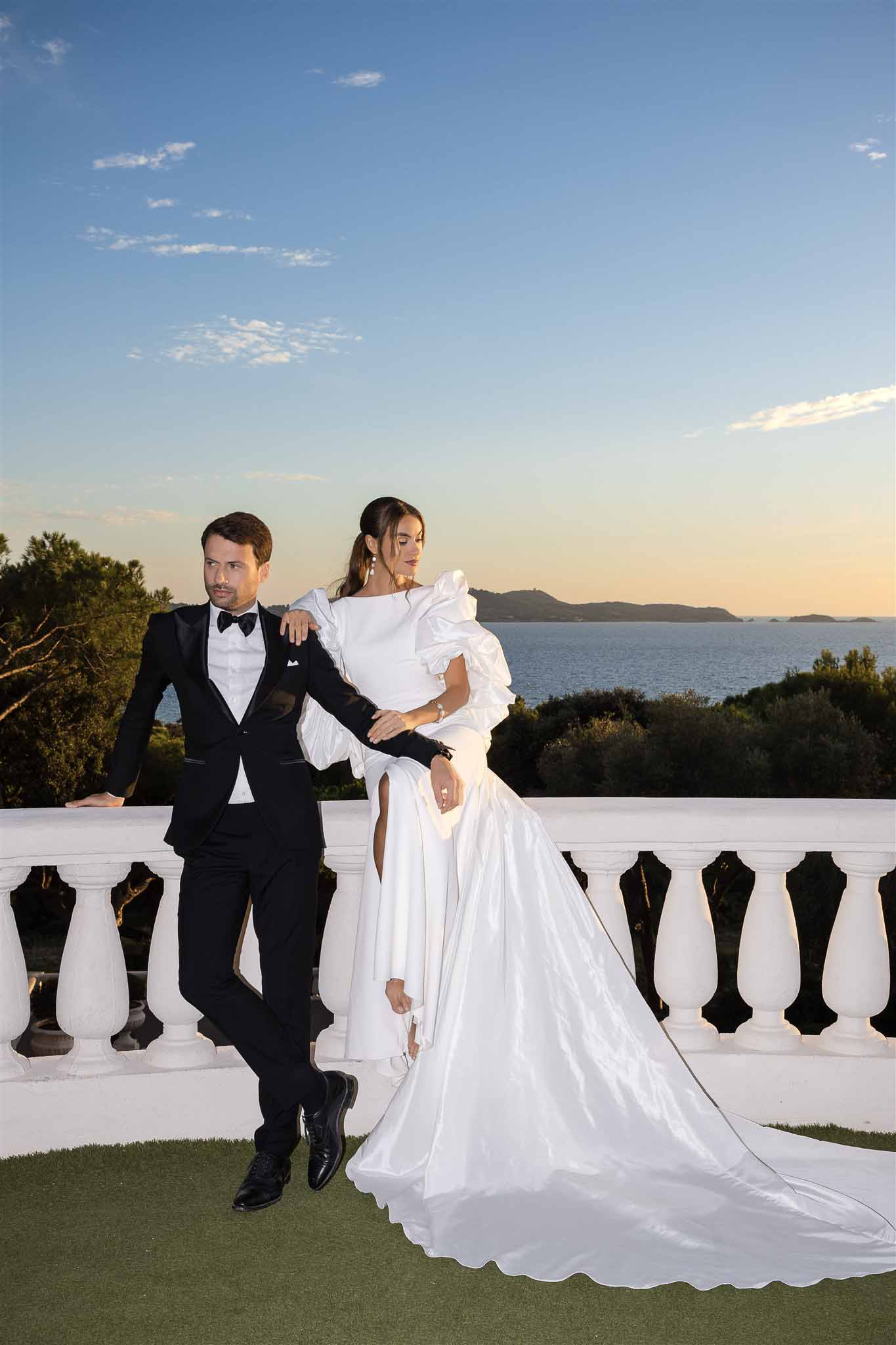Couple on coastal terrace at golden hour, bride in ruffle-sleeve gown with cathedral train, editorial pose