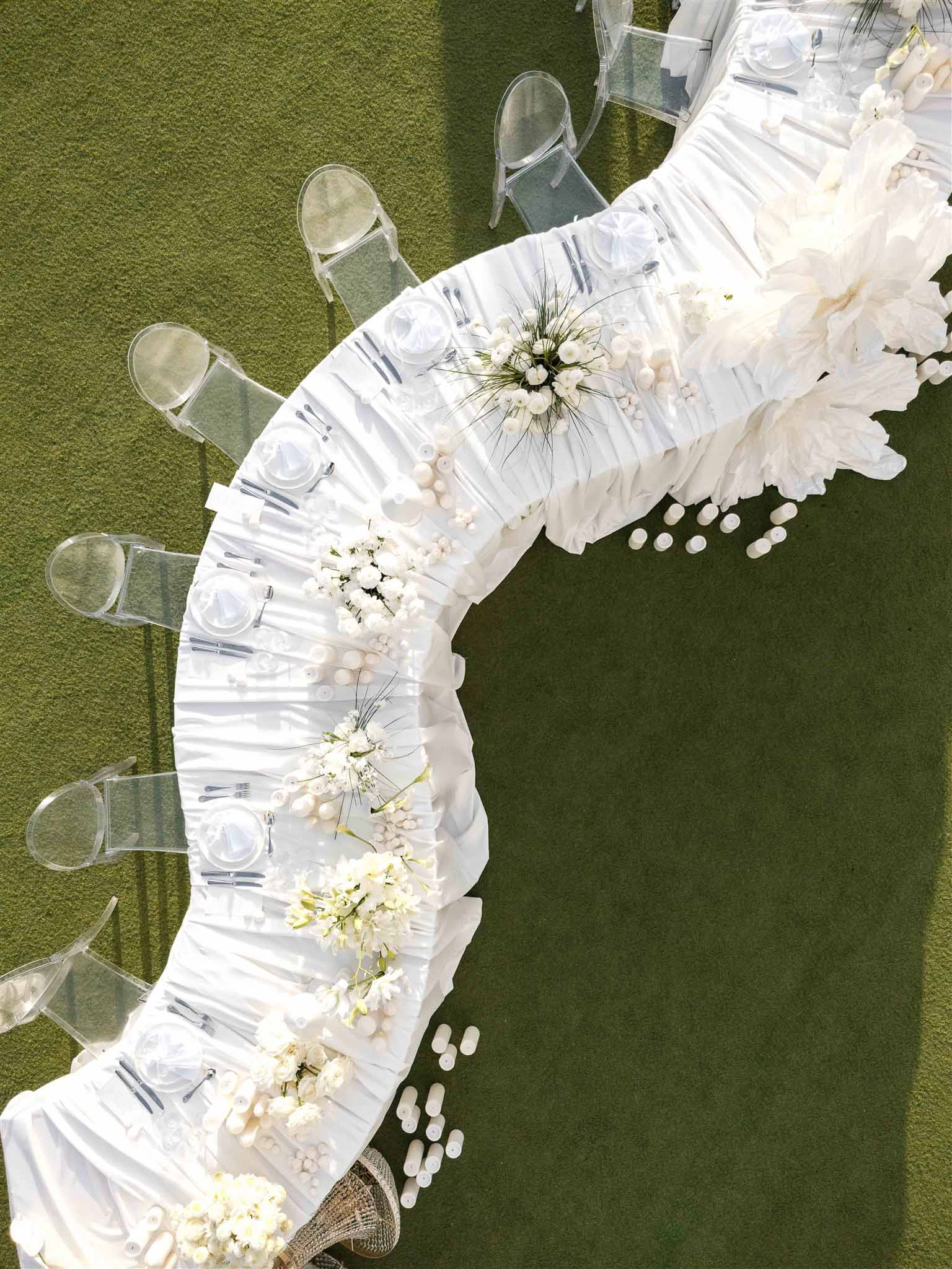 Aerial view of serpentine wedding reception table with all-white floral centerpieces and ghost chairs on lawn