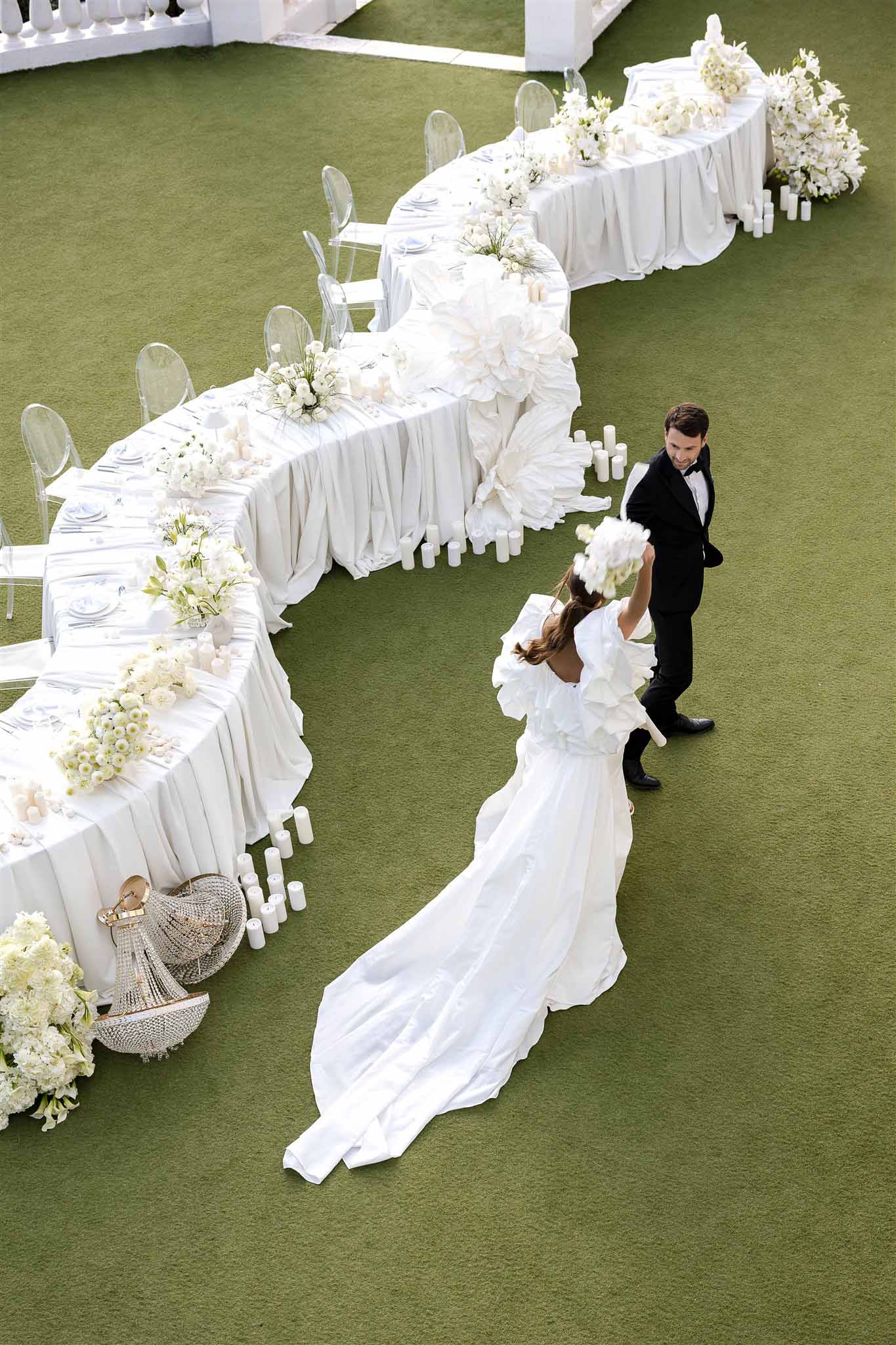 Aerial view of bride and groom beside sinuous white banquet table with ghost chairs and all-white floral installations