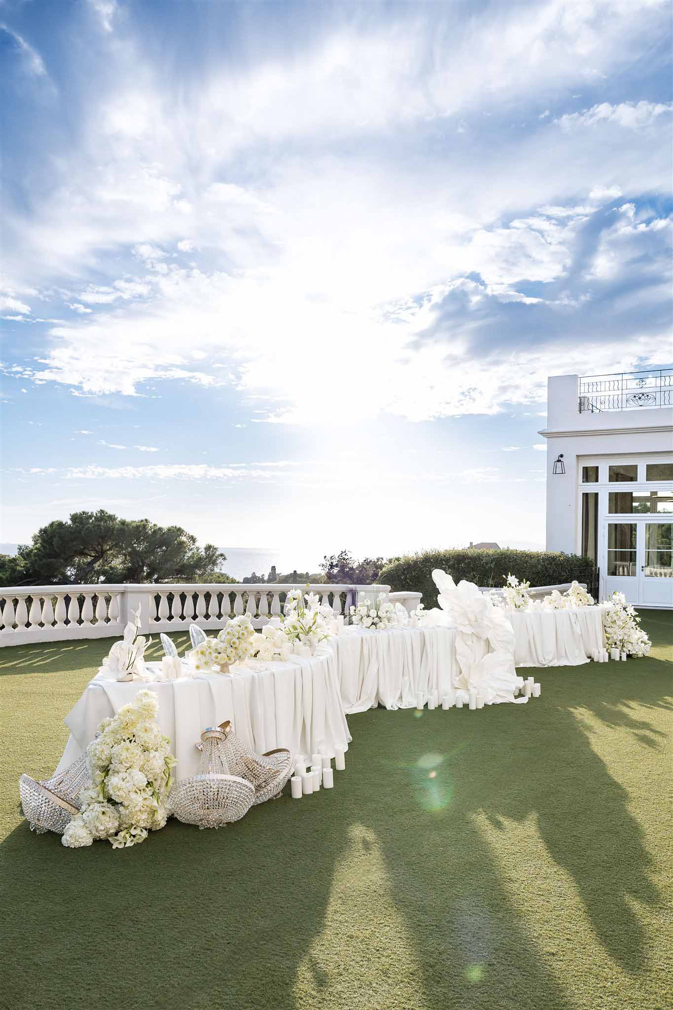 All-white reception table with hydrangeas, pillar candles, and crystal chandeliers on villa lawn with sea views
