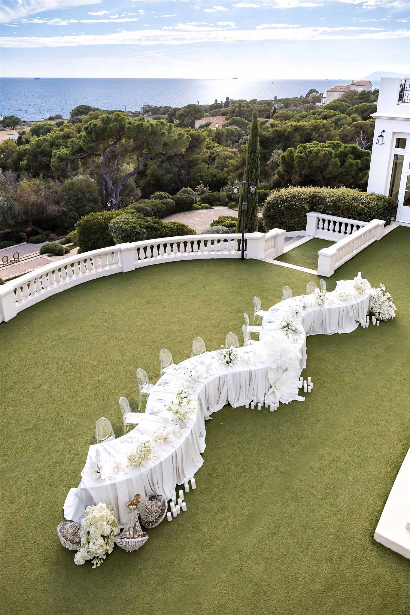 Aerial serpentine white table with ghost chairs, hydrangea centerpieces, and rattan lanterns overlooking sea