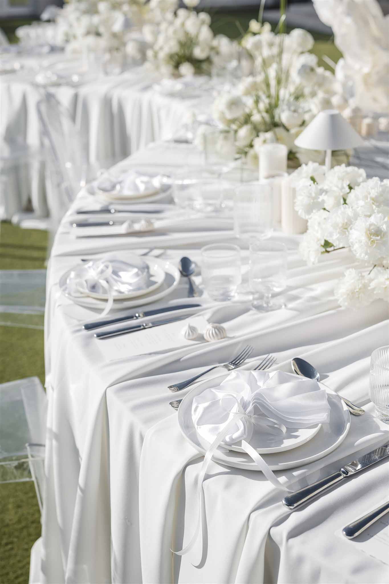 All-white banquet table with peony centerpiece, meringue accents, and ghost chairs on satin cloth